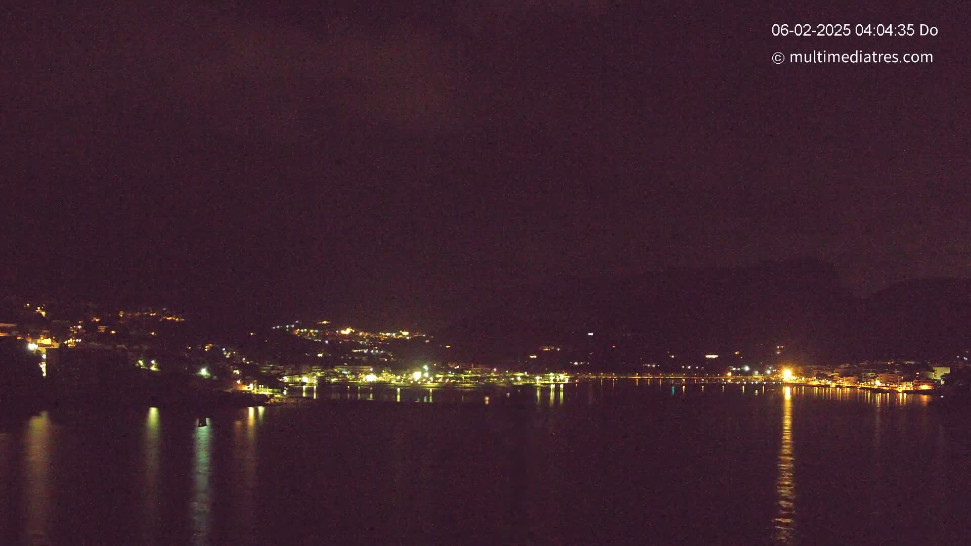 A nighttime view of a coastal town illuminated by lights, reflected on a calm body of water under a dark sky.