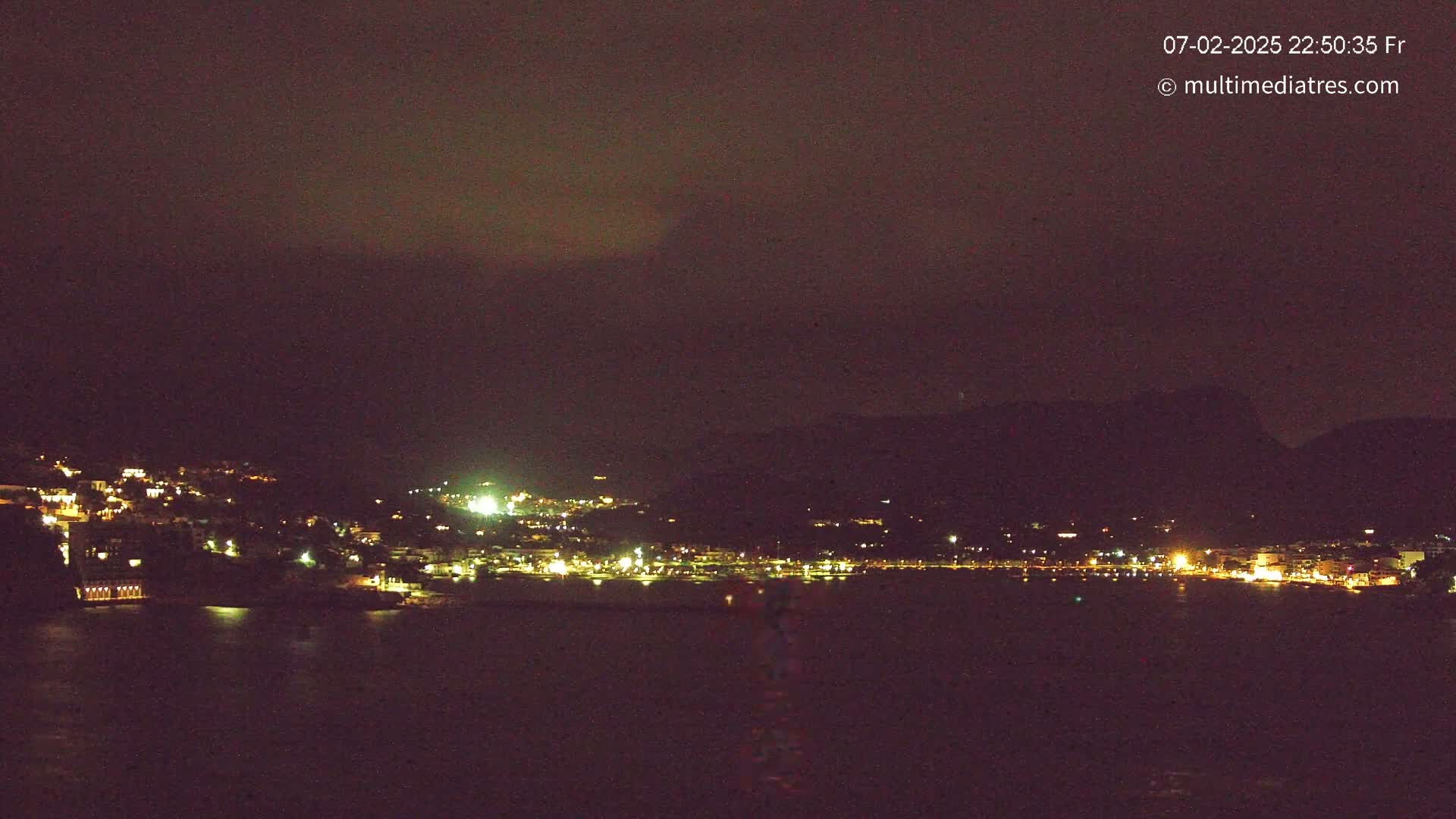 A nighttime, coastal town with many lights is visible across a dark body of water, under a mostly dark sky.