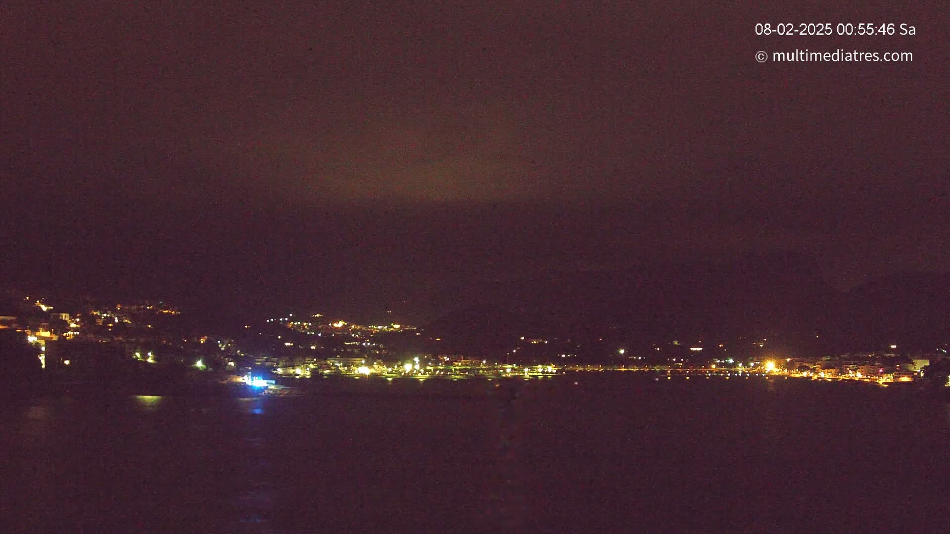 A nighttime coastal town with scattered lights is visible across a dark body of water under a dark, cloudy sky.