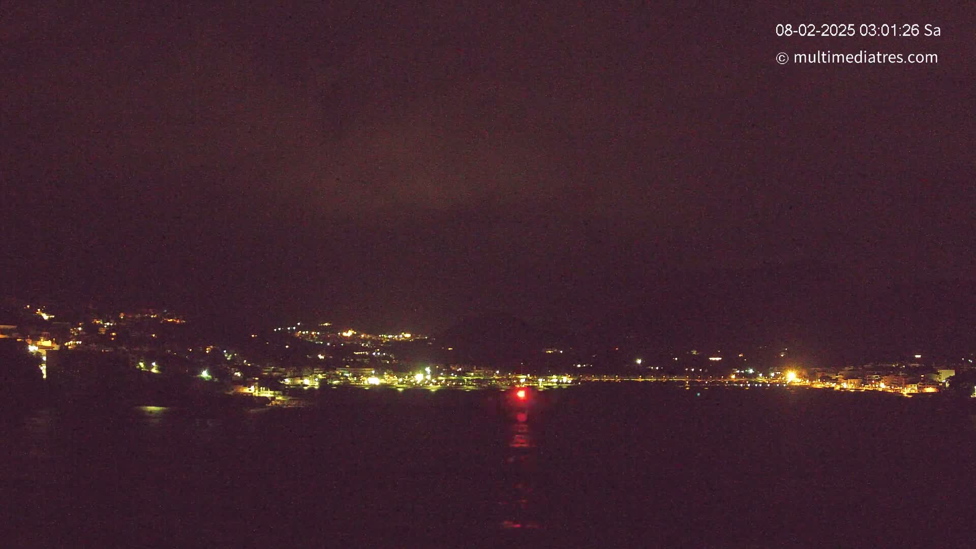 A nighttime, long-range view of a town's lights spread across a hillside next to a dark body of water, under a dark, mostly clear sky.