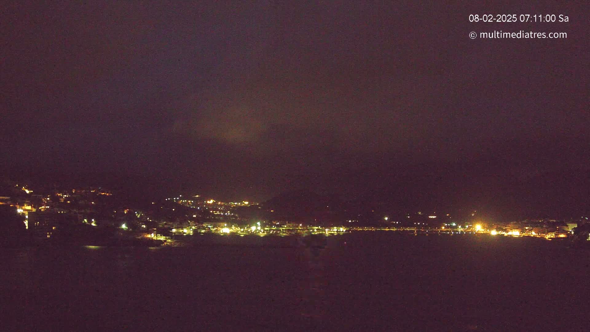 A nighttime long shot shows a coastal town with scattered lights under a dark, overcast sky.