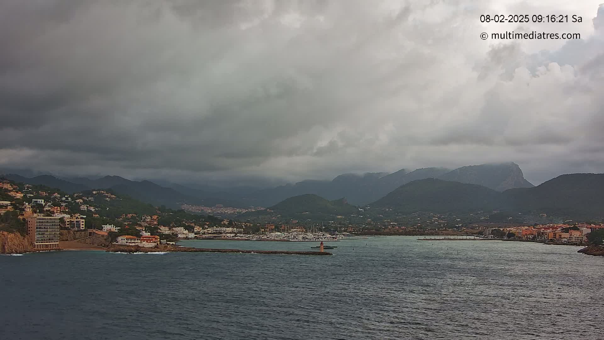 A coastal town nestled between dark, overcast skies and a range of mountains, with a harbor filled with boats.