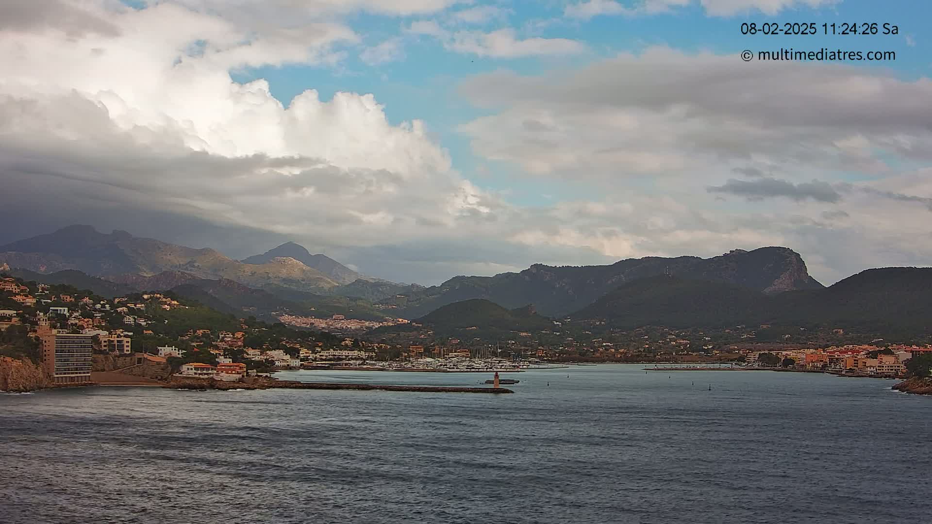 A partly cloudy sky overlooks a coastal town nestled between dark mountains and a calm bay filled with boats.