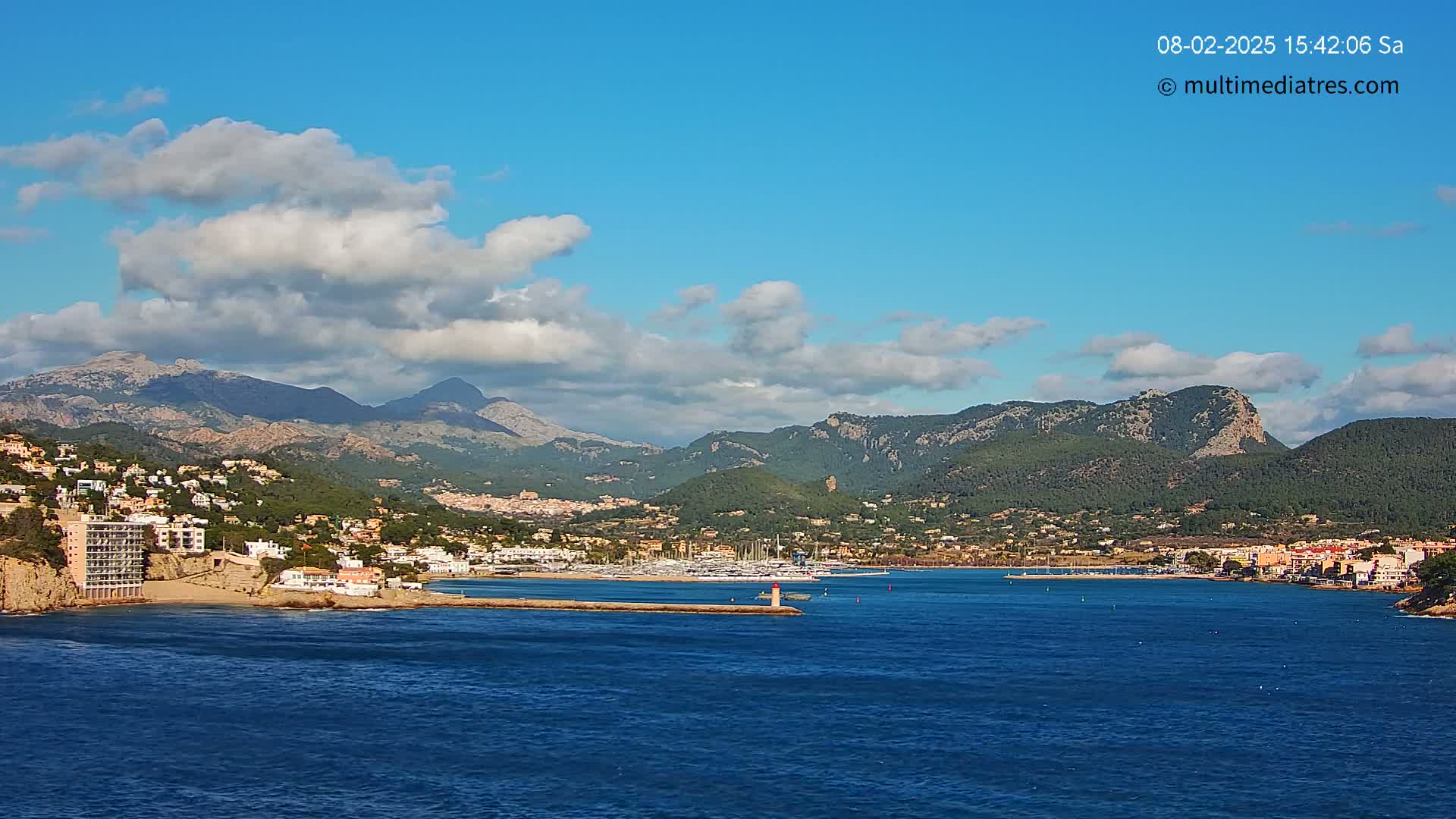 A partly cloudy blue sky overlooks a coastal town nestled between mountains and a calm blue sea.