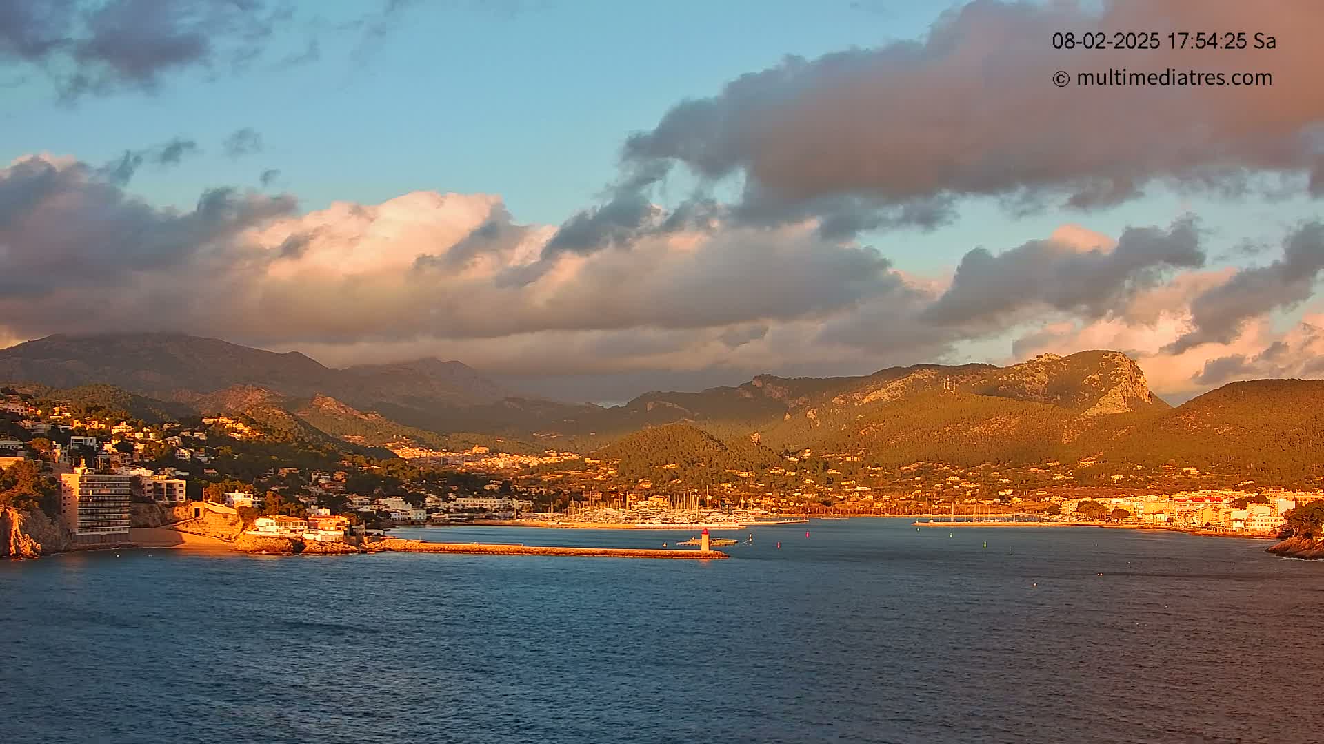 A coastal town nestled against sunlit mountains under a partly cloudy sunset sky.