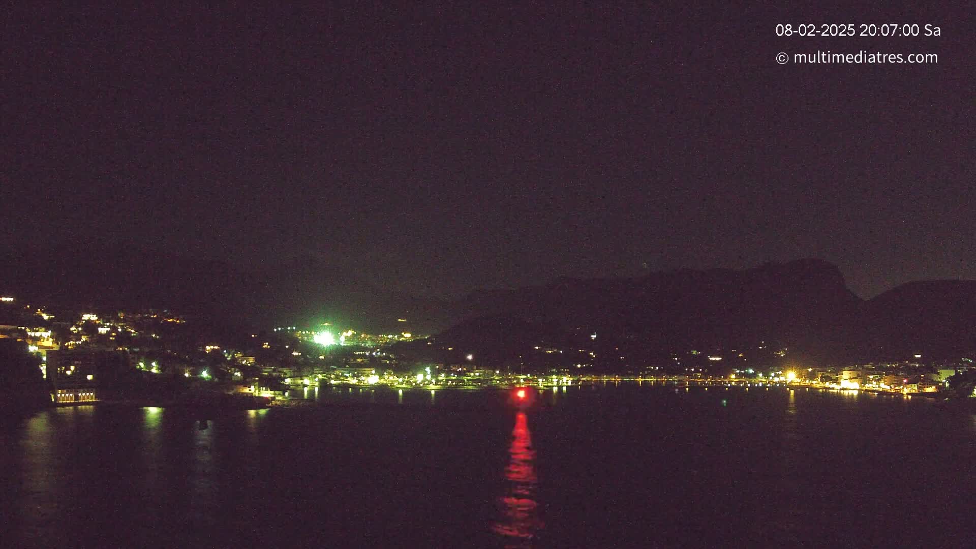 A nighttime view of a dark, calm body of water with a town illuminated by lights nestled between dark hills under a clear night sky.