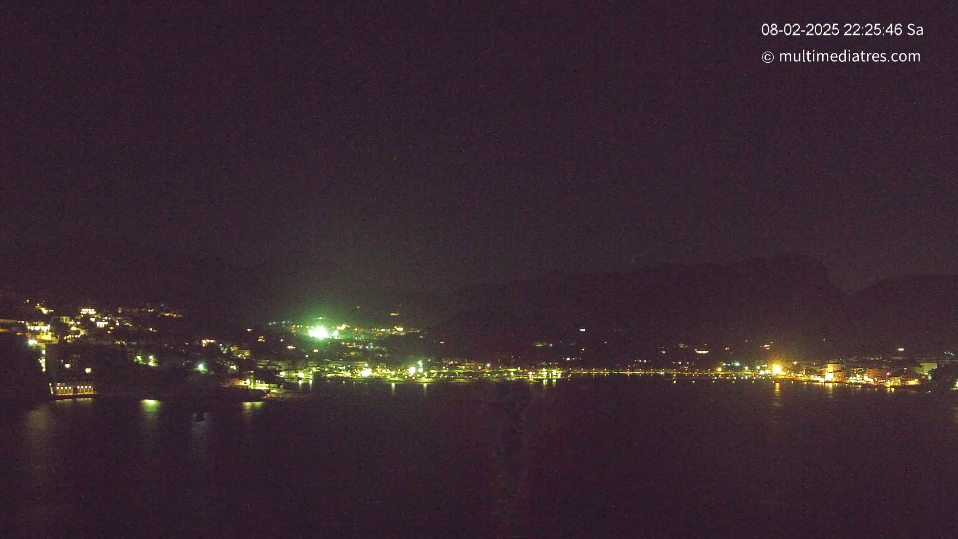 A nighttime view of a coastal town with its lights reflecting on a dark body of water under a clear, dark sky.