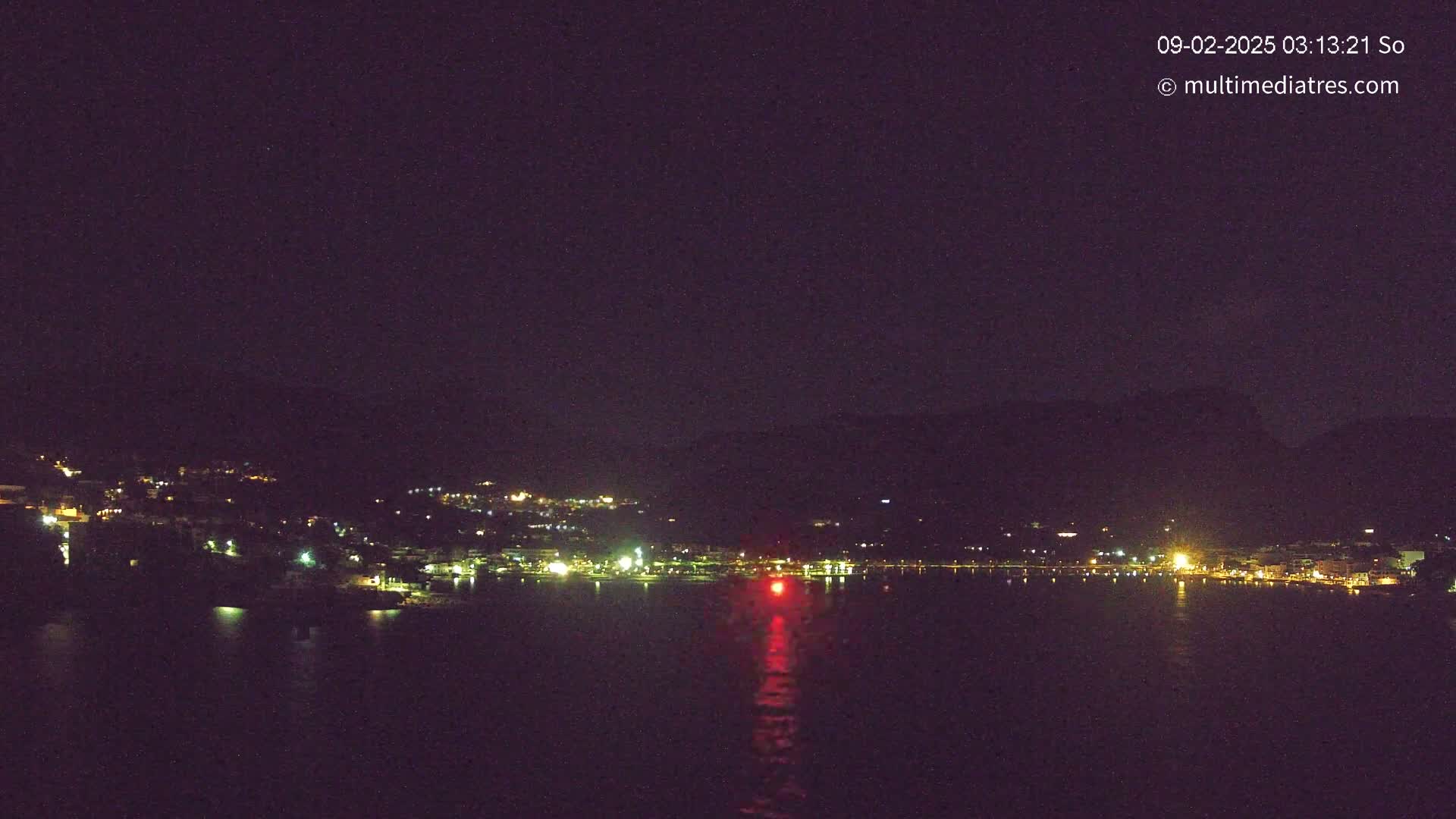 A nighttime view of a town's lights across a dark body of water, under a clear, dark sky.