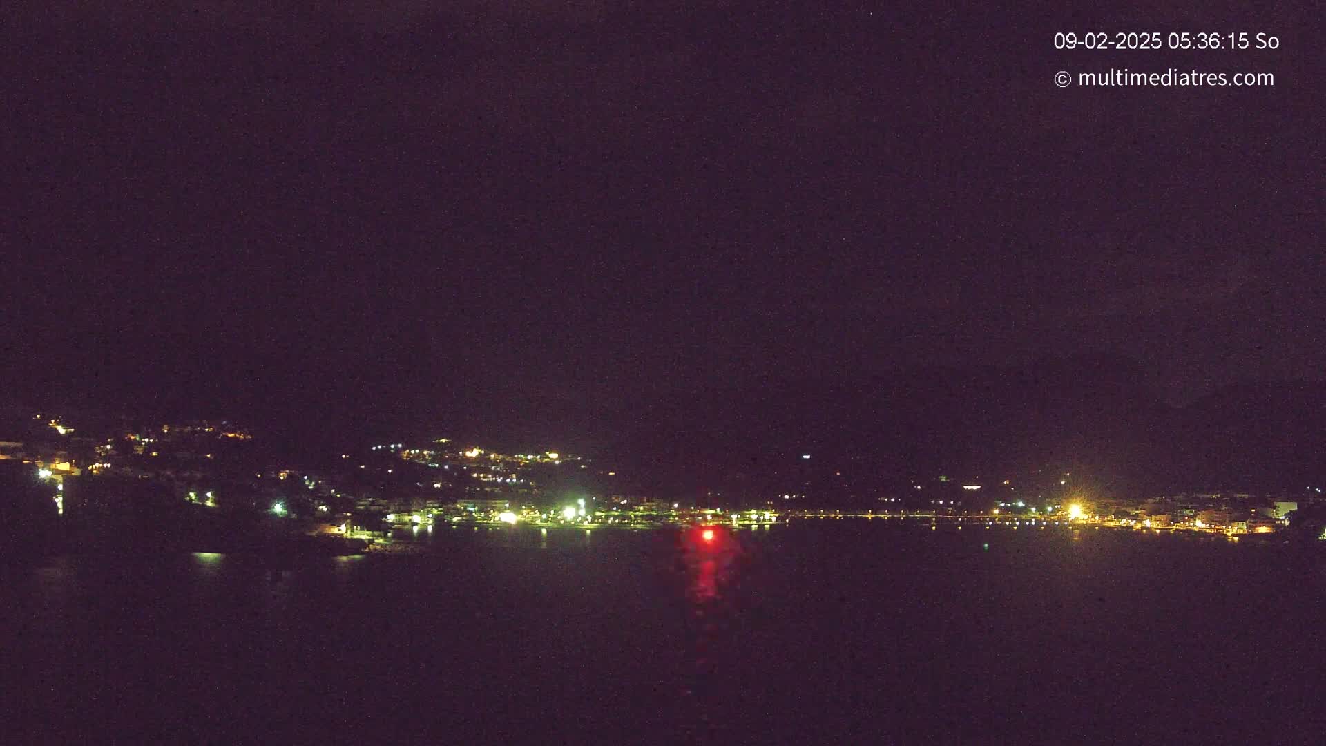 A nighttime view of a town's lights along a dark body of water under a clear, dark sky.
