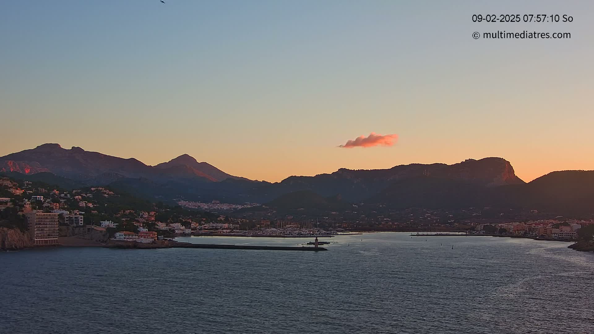A coastal town nestled between mountains at sunset, with a calm body of water in the foreground and a single pink cloud in the sky.