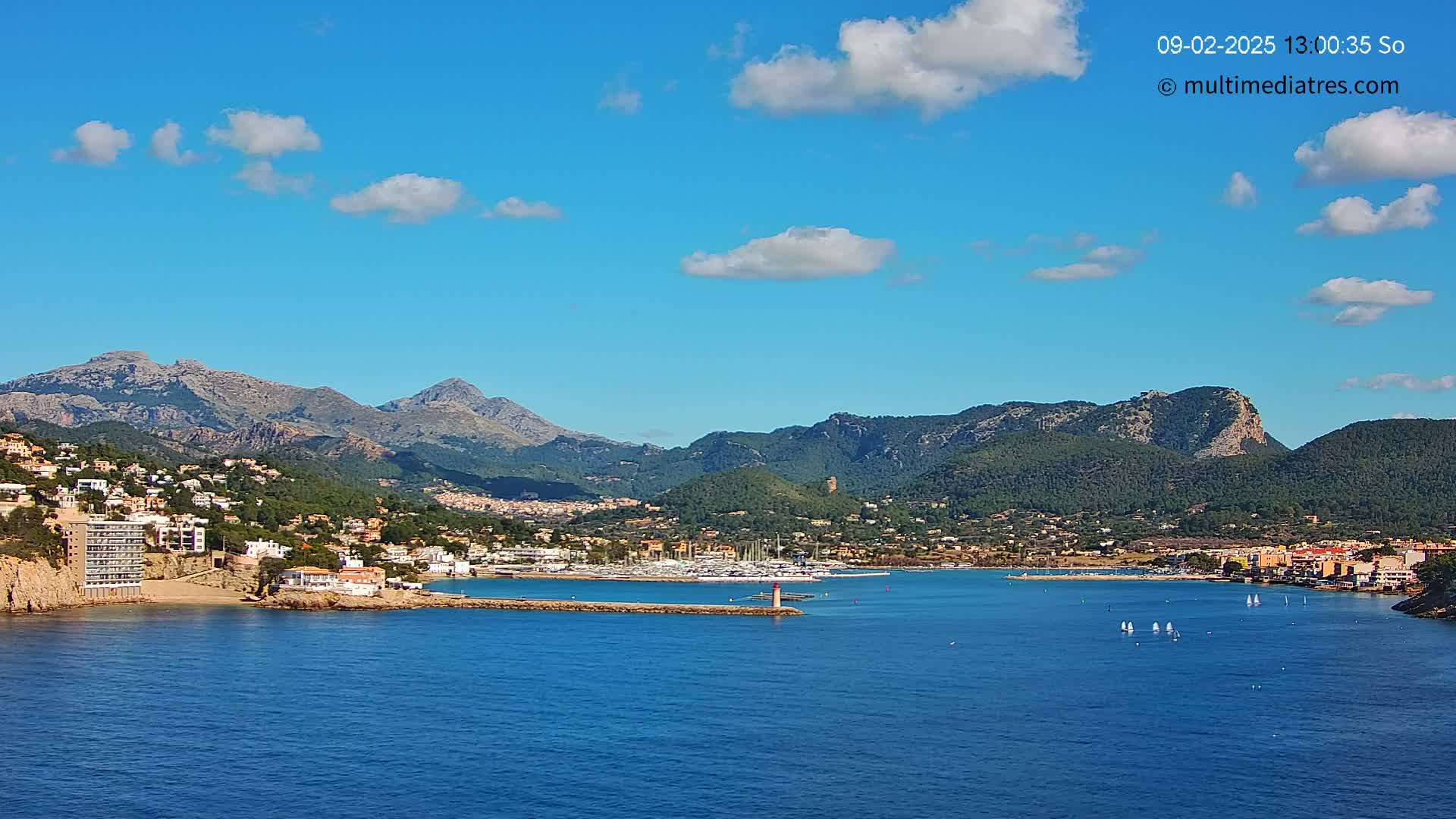 A coastal town nestled between mountains, with a marina filled with boats, under a mostly sunny sky with a few scattered clouds.