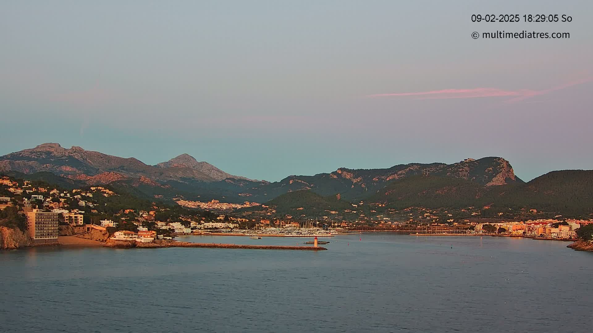 A coastal town nestled between mountains at dusk under a pale pink and blue sky, with a calm harbor containing numerous boats.