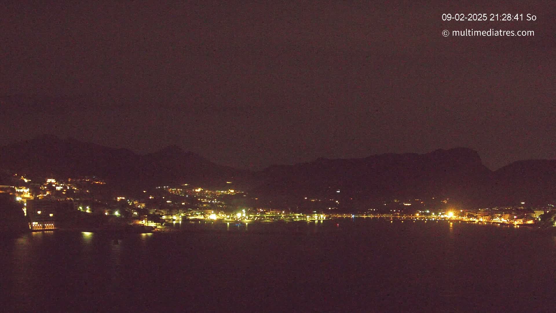 A nighttime view of a coastal town nestled between dark mountains, with its lights reflected on a calm body of water under a clear, dark sky.
