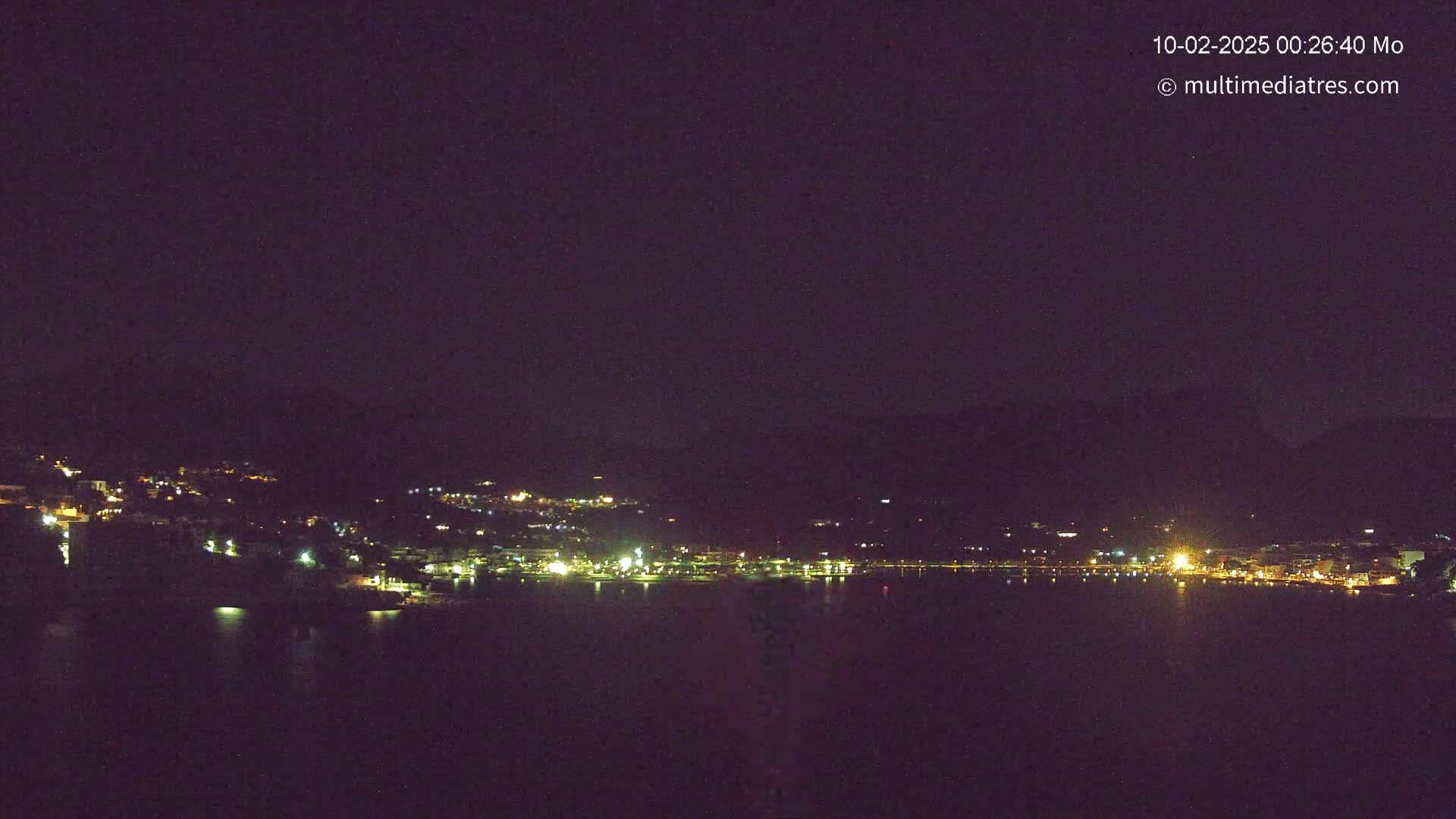 A nighttime view of a town's lights nestled along a dark body of water, under a clear, starless sky.