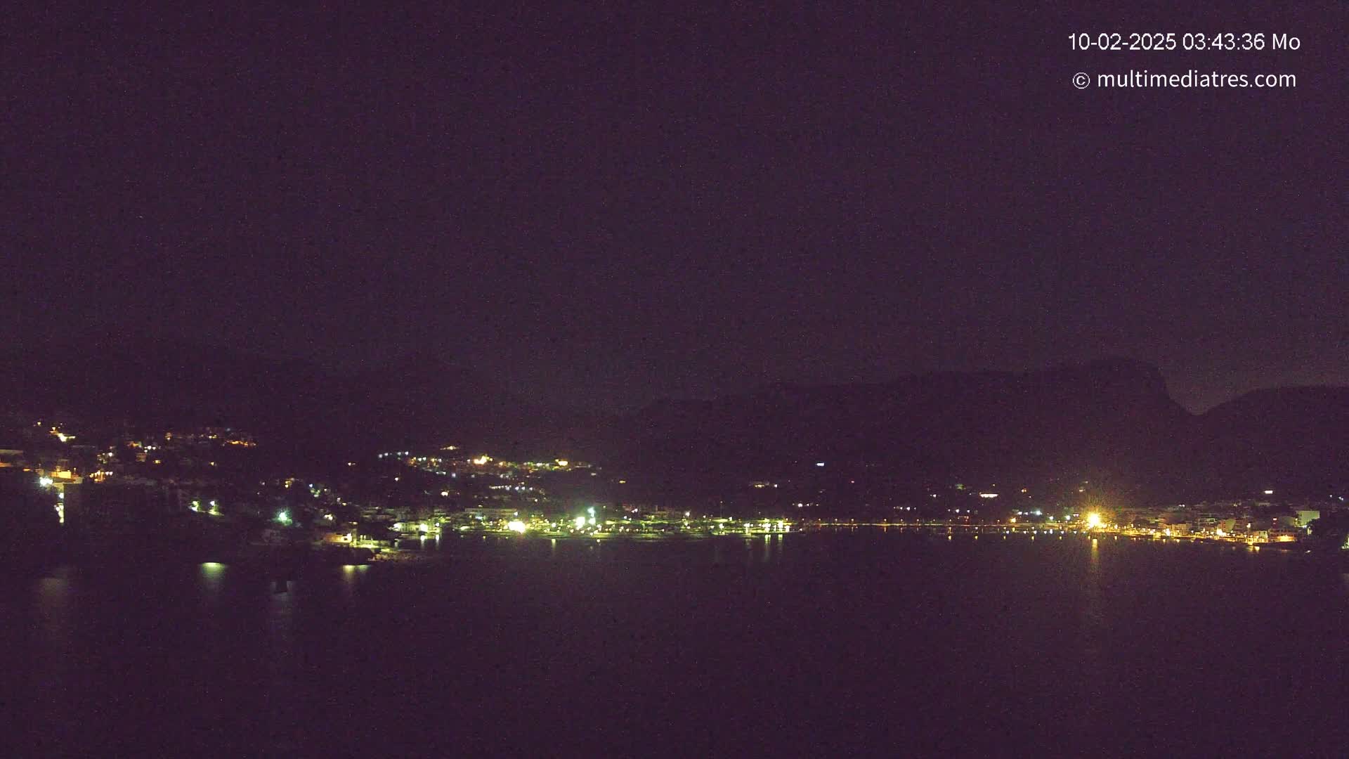 A nighttime long shot of a town situated on a body of water, nestled between hills under a dark sky.