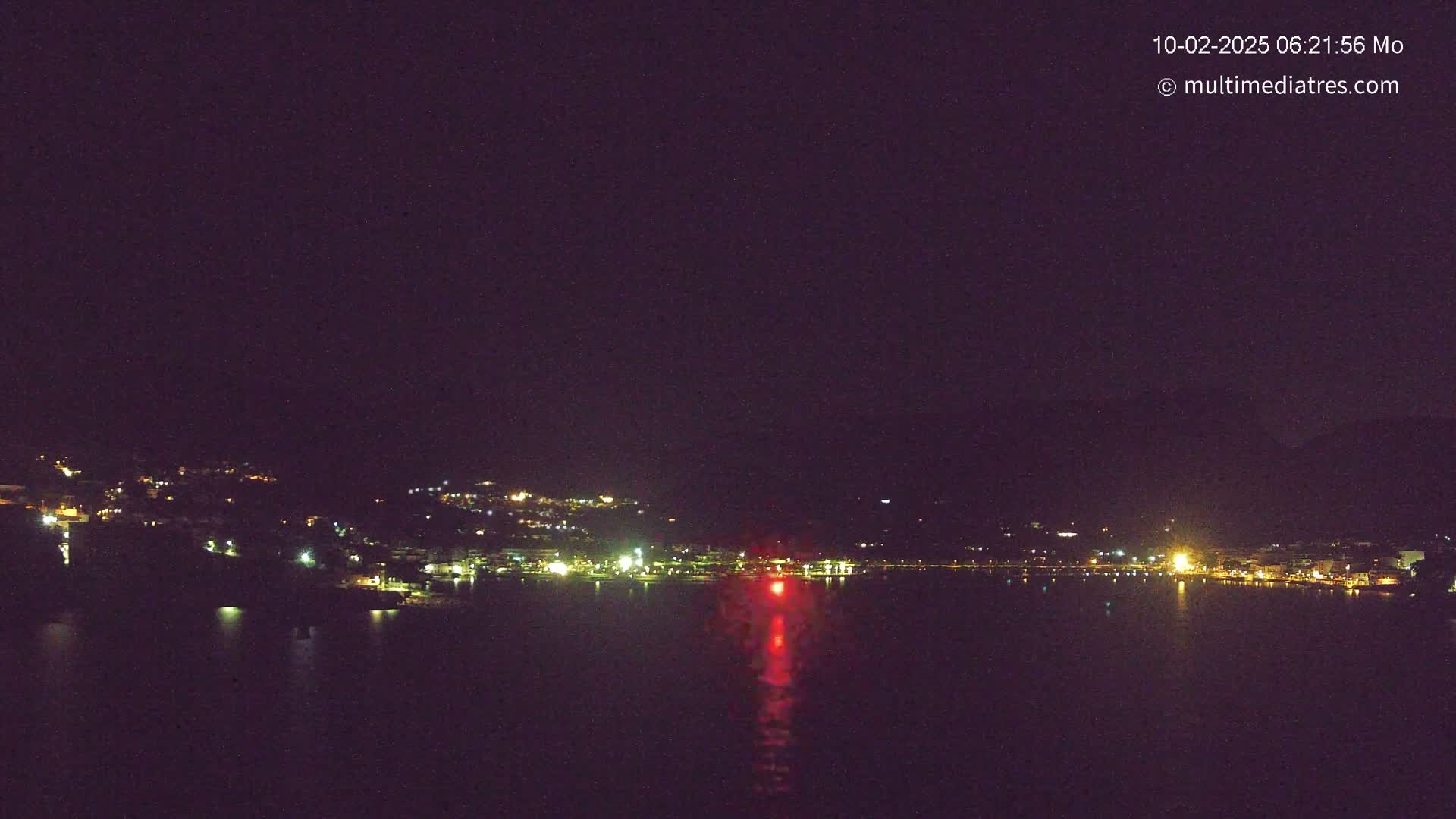 A nighttime view of a town's lights along a dark body of water, with a single red light reflecting on the water's surface under a clear, dark sky.