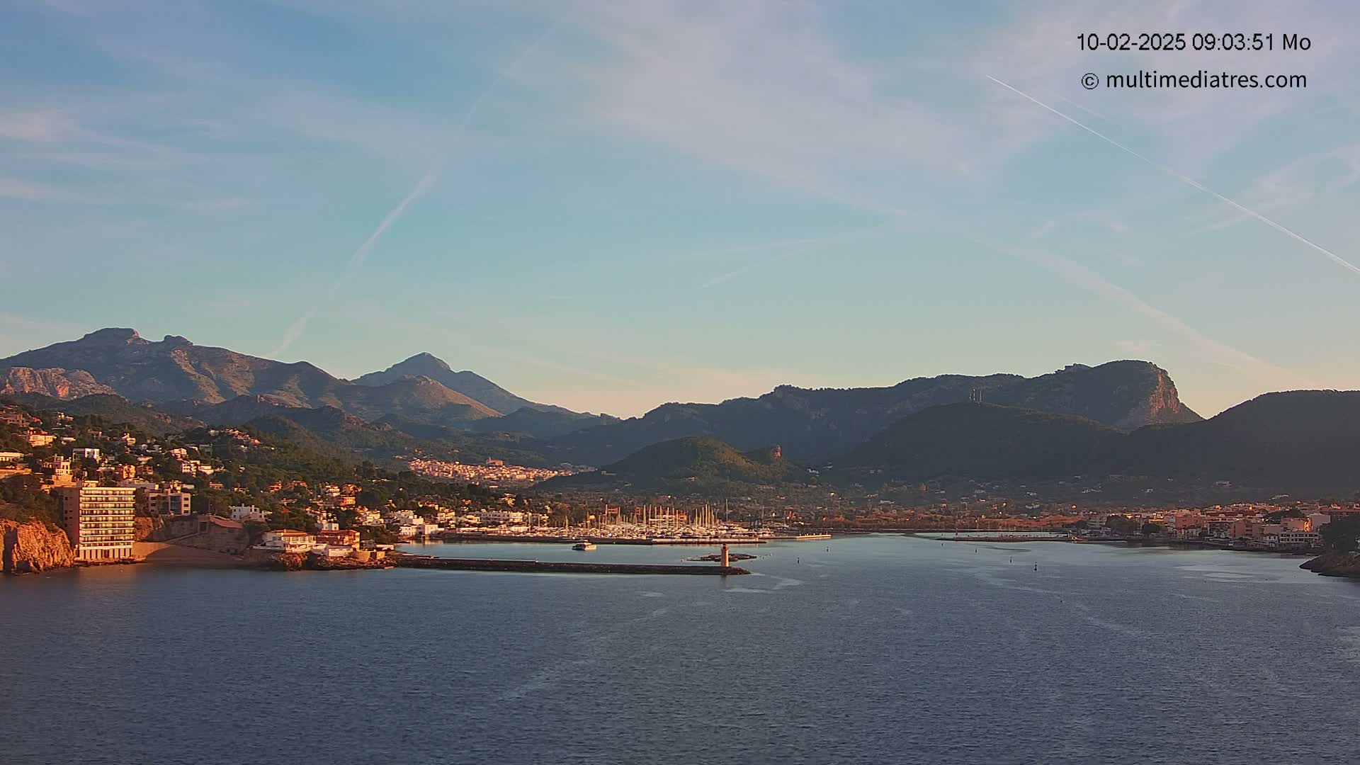 A coastal town nestled between mountains under a clear, light blue sky, with a calm body of water in the foreground containing a marina.