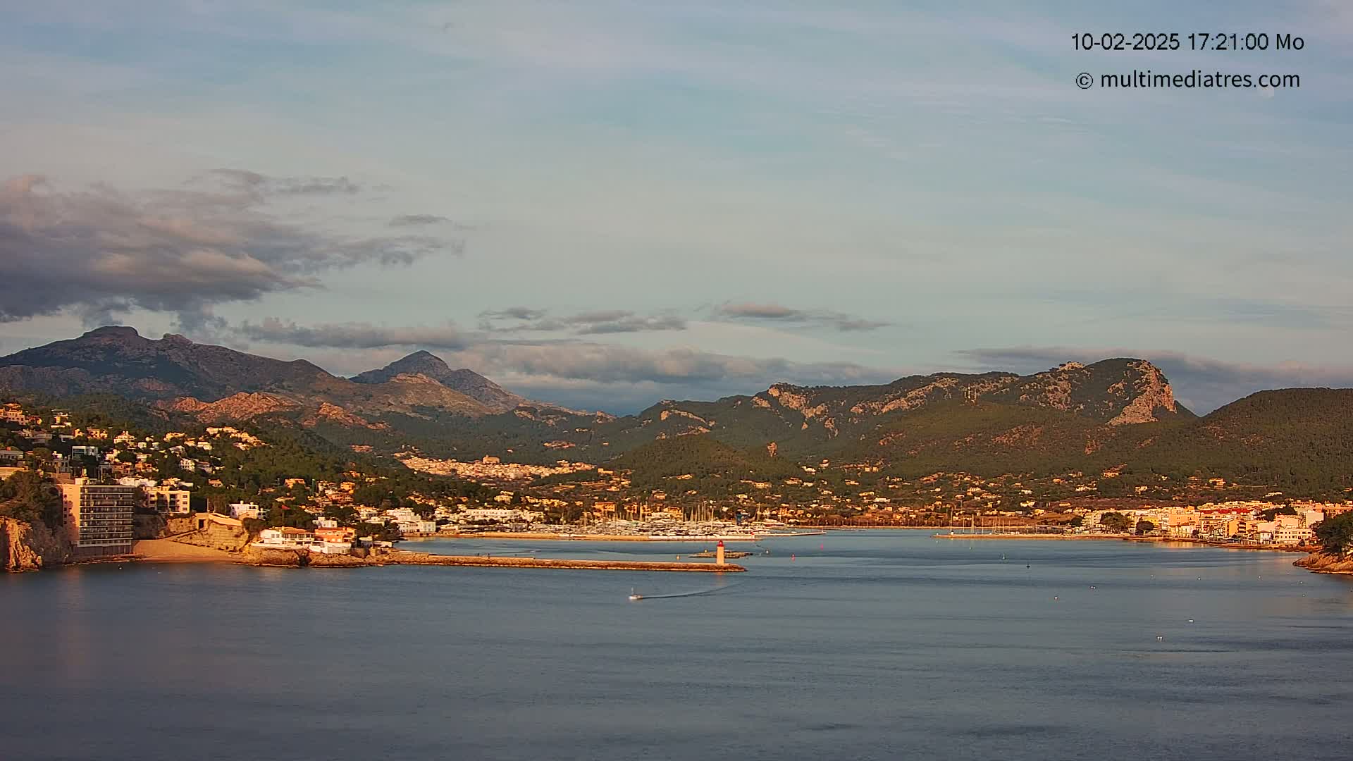 A coastal town nestled at the base of a mountain range, with a calm bay filled with boats under a partly cloudy sky.