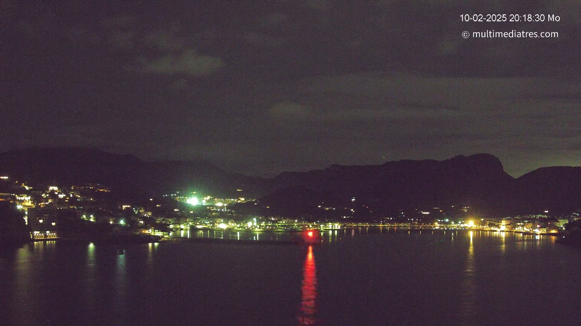 A nighttime view of a city nestled between dark hills and a calm body of water, under a mostly cloudy sky.