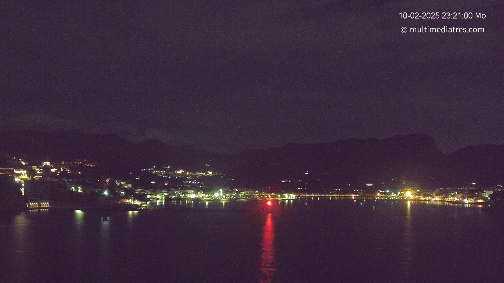 A nighttime view of a town situated along a dark body of water, bordered by mountains under a clear, dark sky.