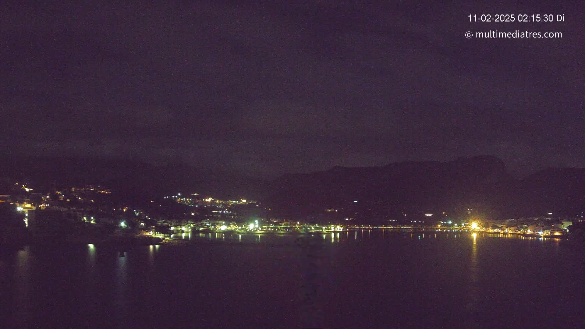 A nighttime view of a town situated on a body of water, nestled against dark hills under a mostly clear, dark sky.