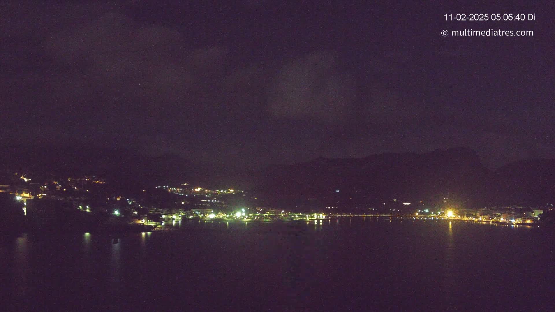 A nighttime view of a town situated on a body of water, nestled against dark hills under a partly cloudy sky.