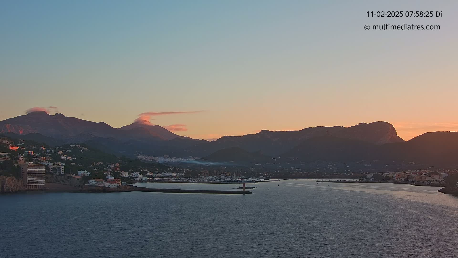 A coastal town nestled between mountains at sunset, with calm waters in the foreground.