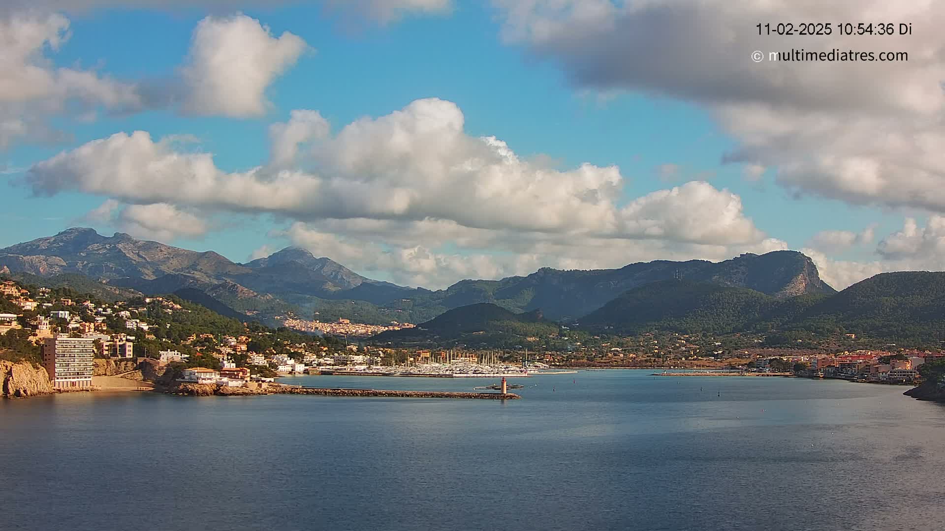 A coastal town nestled between mountains under a partly cloudy sky, with a harbor filled with boats.