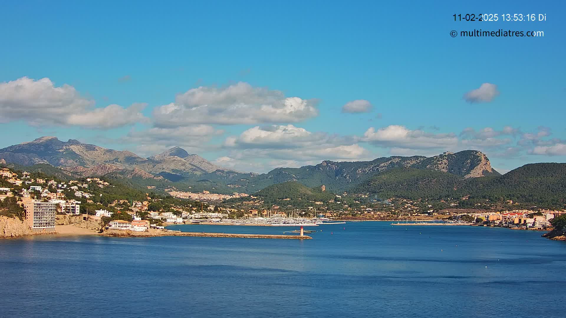 A coastal town nestled against a mountainous backdrop under a mostly sunny sky with scattered clouds, features a calm blue sea with a marina filled with boats.