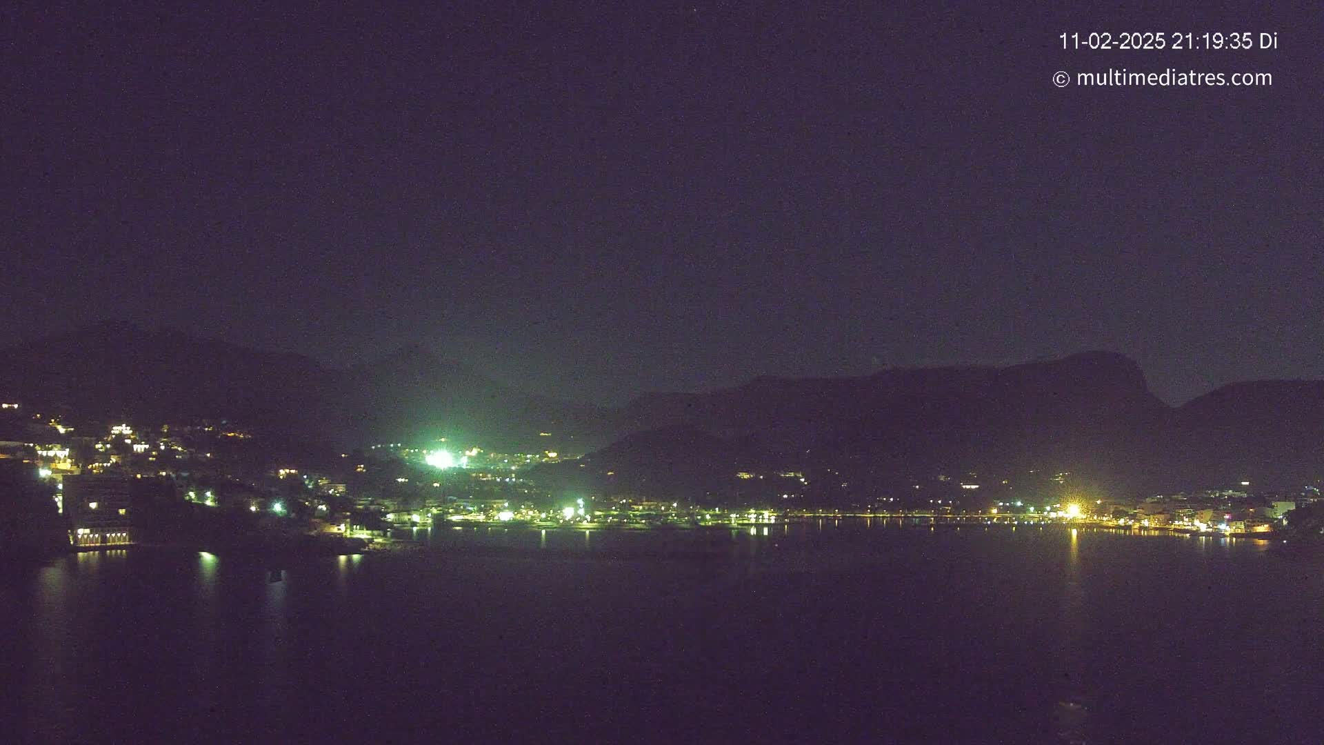 A nighttime view of a town nestled between dark mountains and a calm body of water, under a clear, dark sky.
