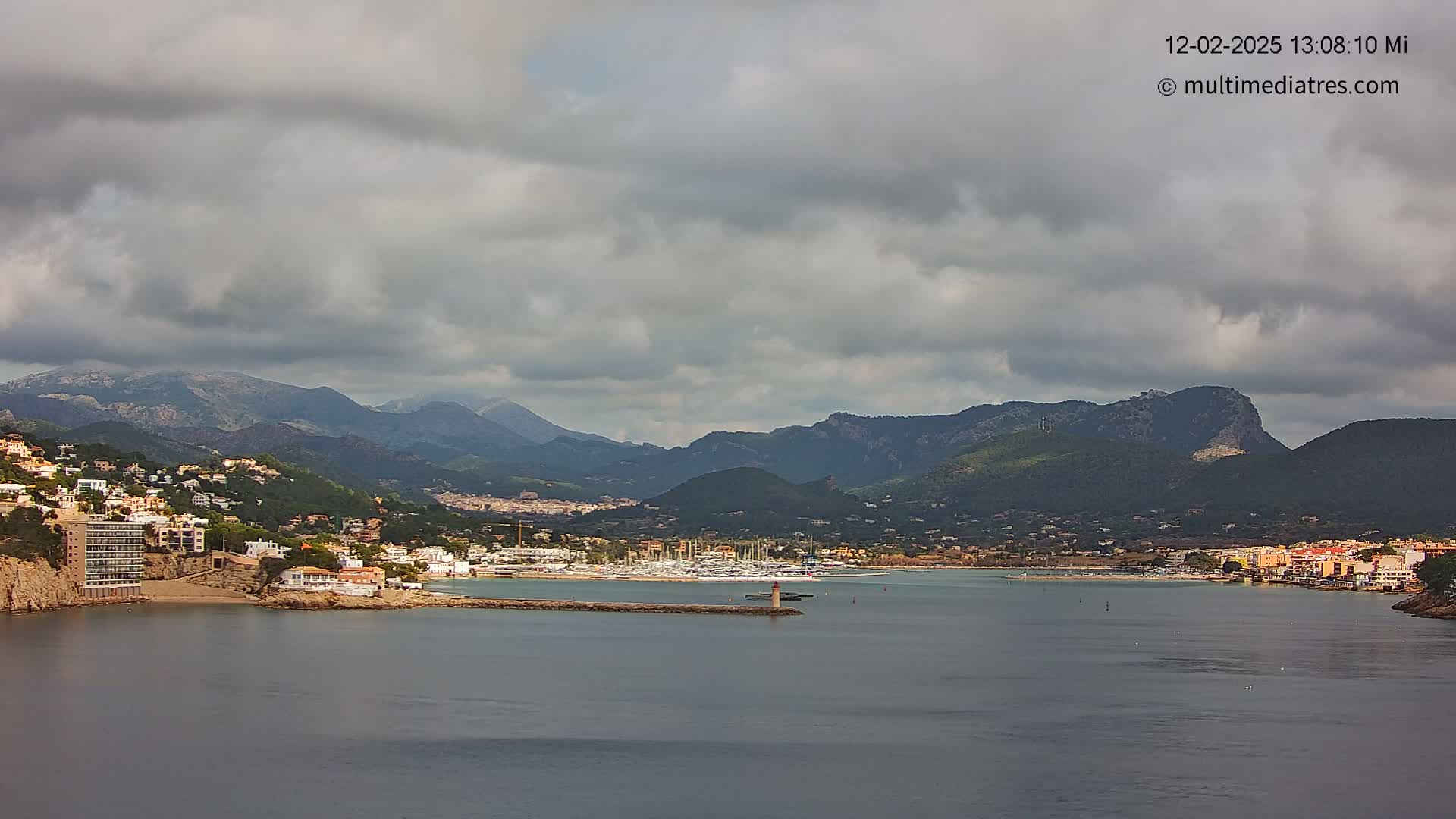 A coastal town nestled between mountains, under a mostly cloudy sky, features a harbor filled with boats.