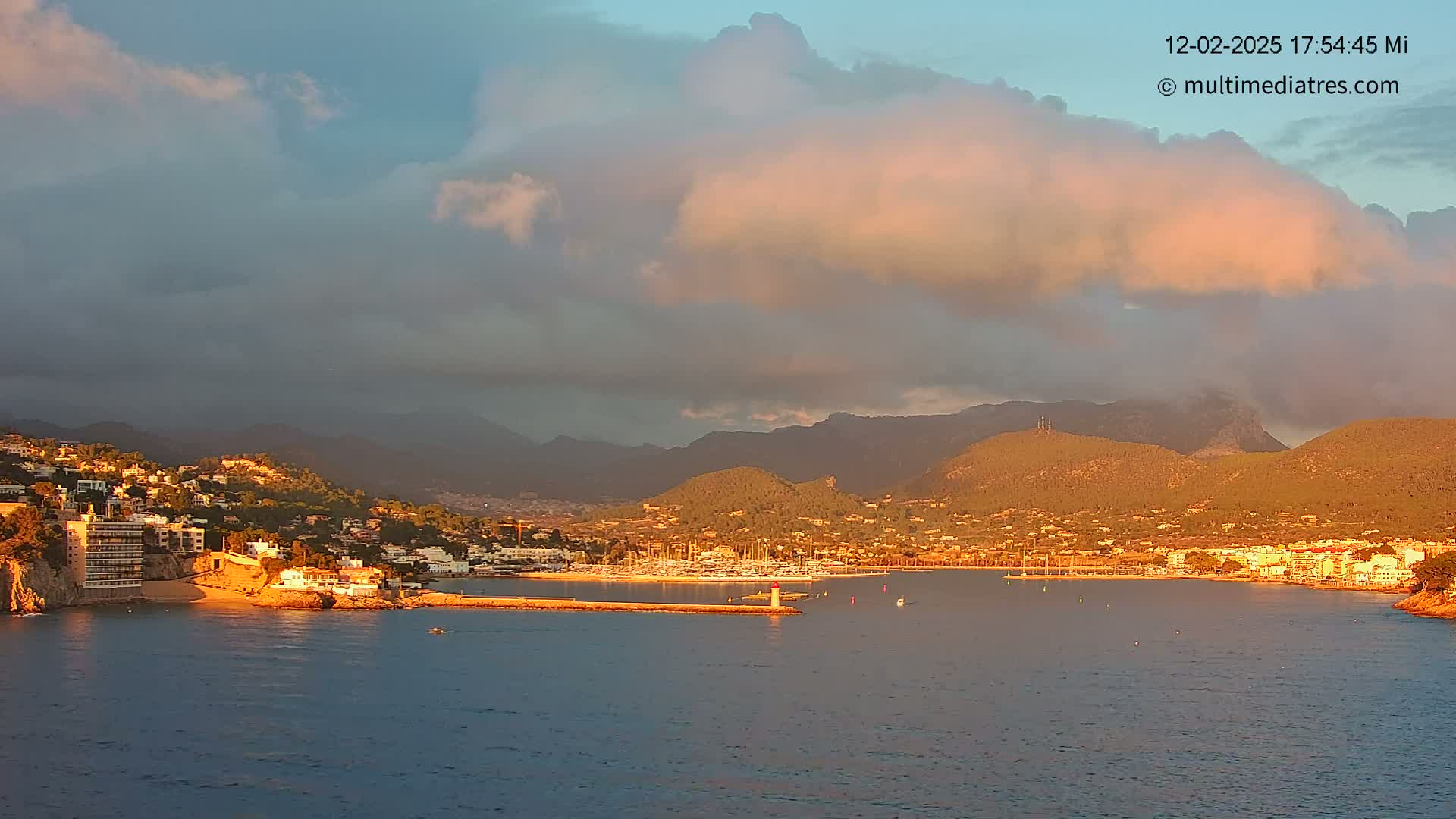 A coastal town nestled against mountains is bathed in the warm glow of the setting sun under a partly cloudy sky.