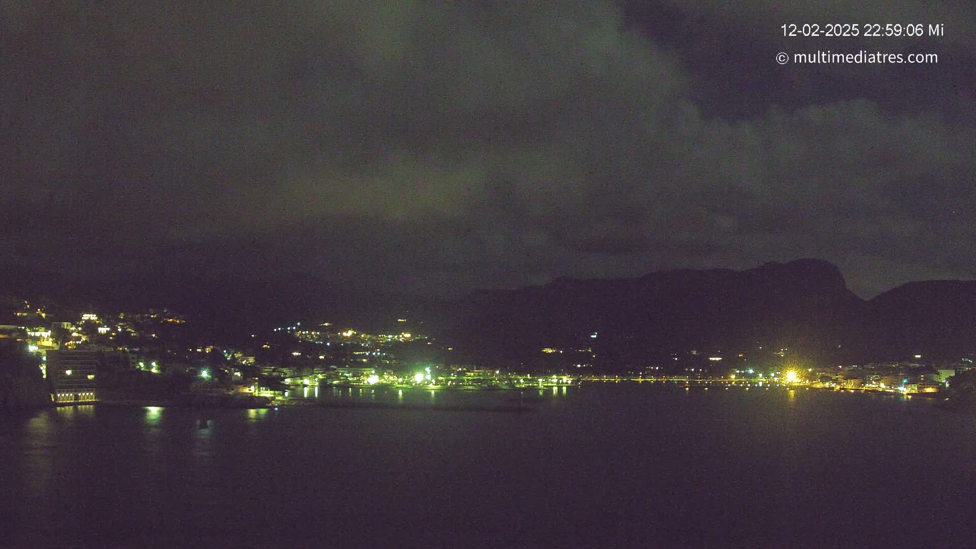 A nighttime view of a coastal town illuminated by scattered lights, under a dark, cloudy sky.