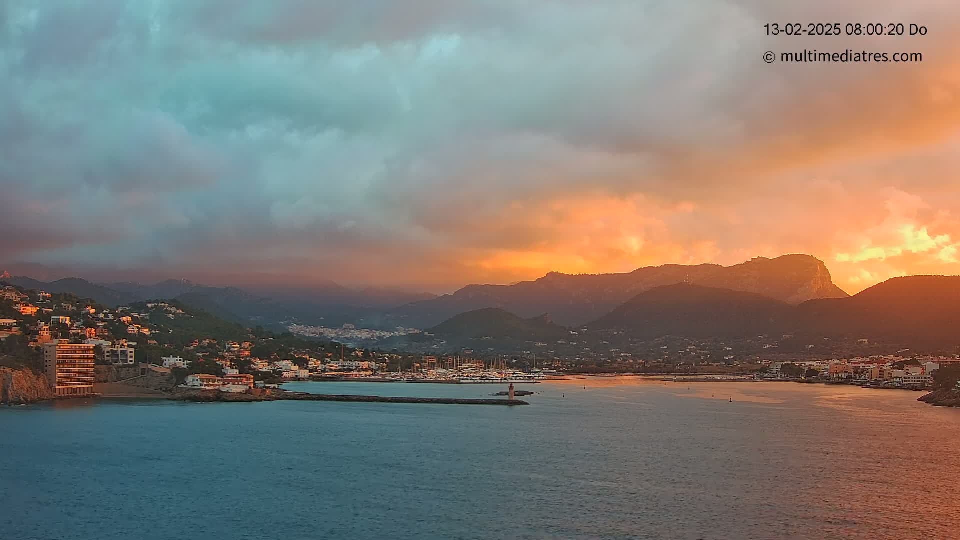 A coastal town nestled between mountains is bathed in the warm glow of a sunset under a partly cloudy sky.