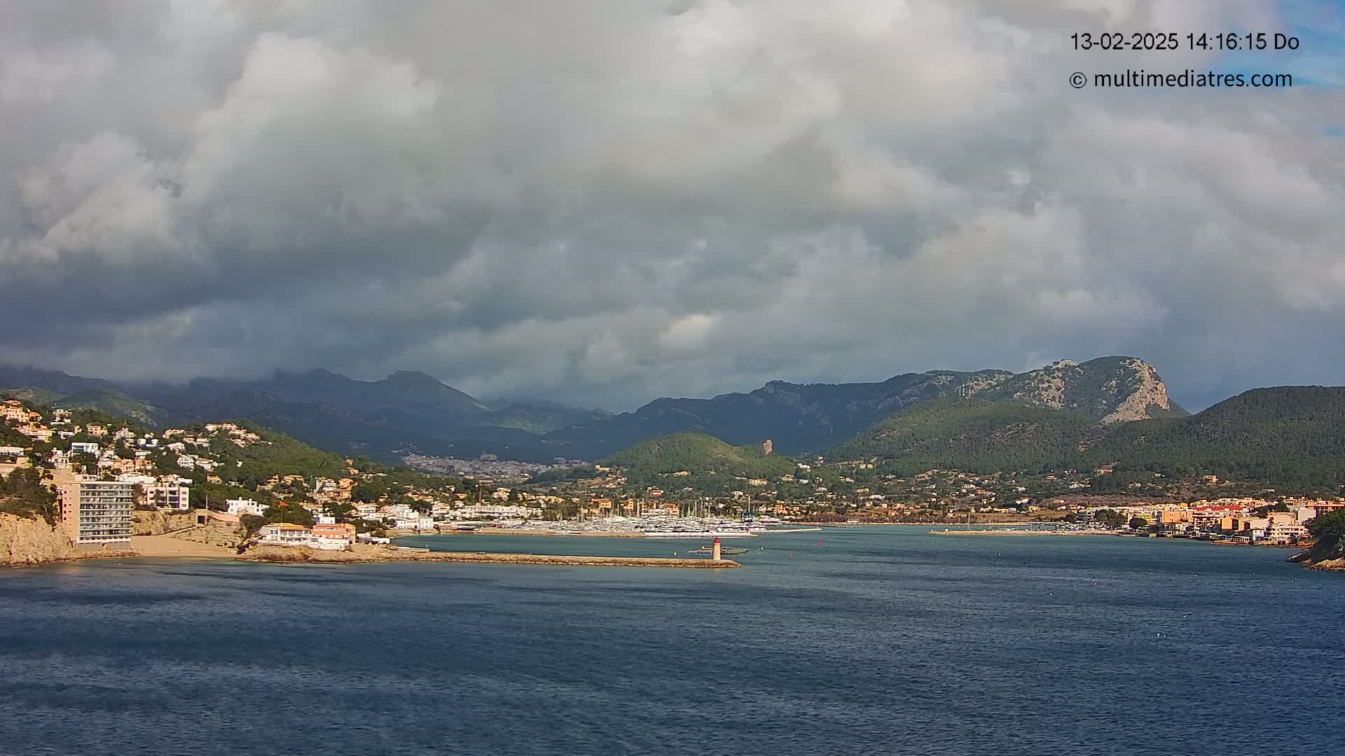 A coastal town nestled in a bay, surrounded by green hills and mountains under a partly cloudy sky.