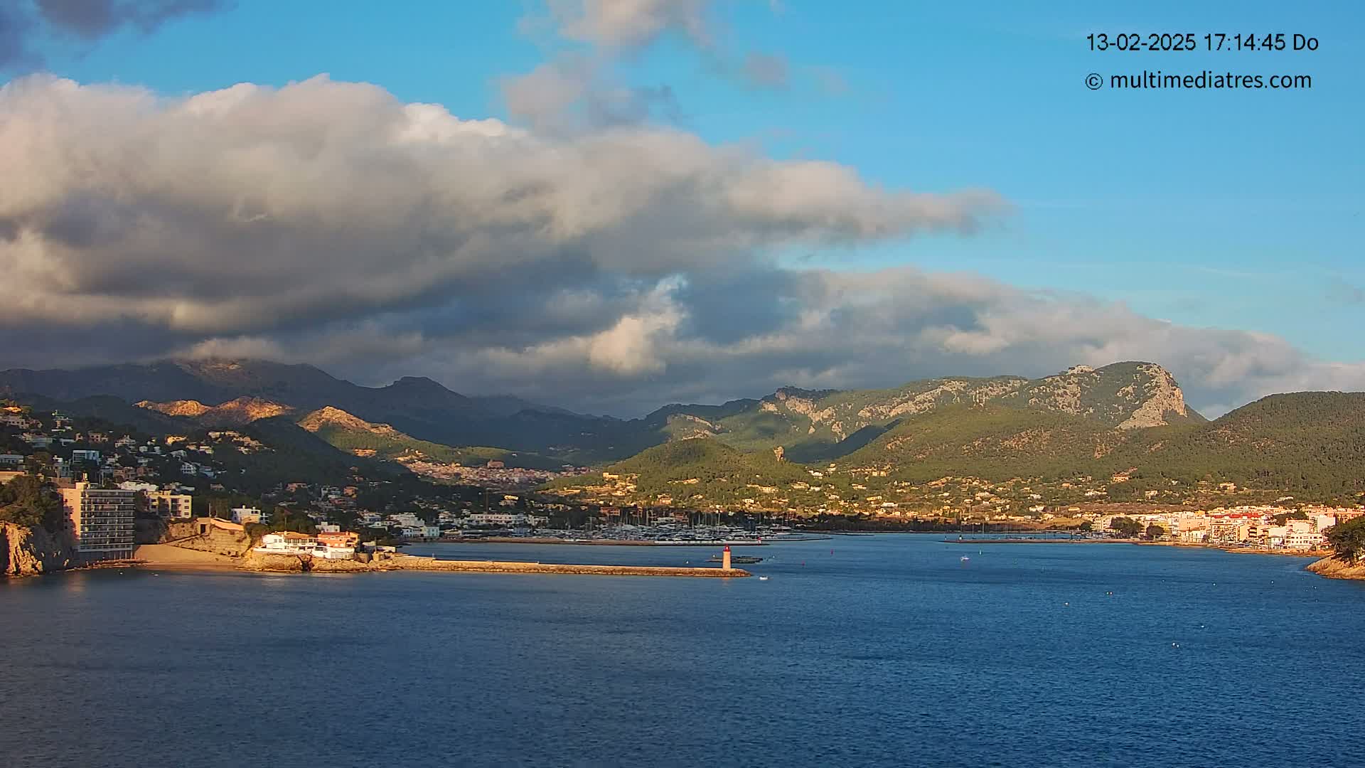 A coastal town nestled between mountains under a partly cloudy sky, with a calm body of water featuring a small harbor.