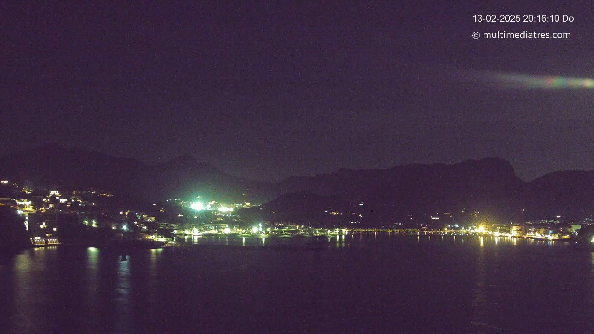 A nighttime view of a coastal town nestled between dark mountains, with its lights reflecting on a calm body of water under a clear, dark sky.