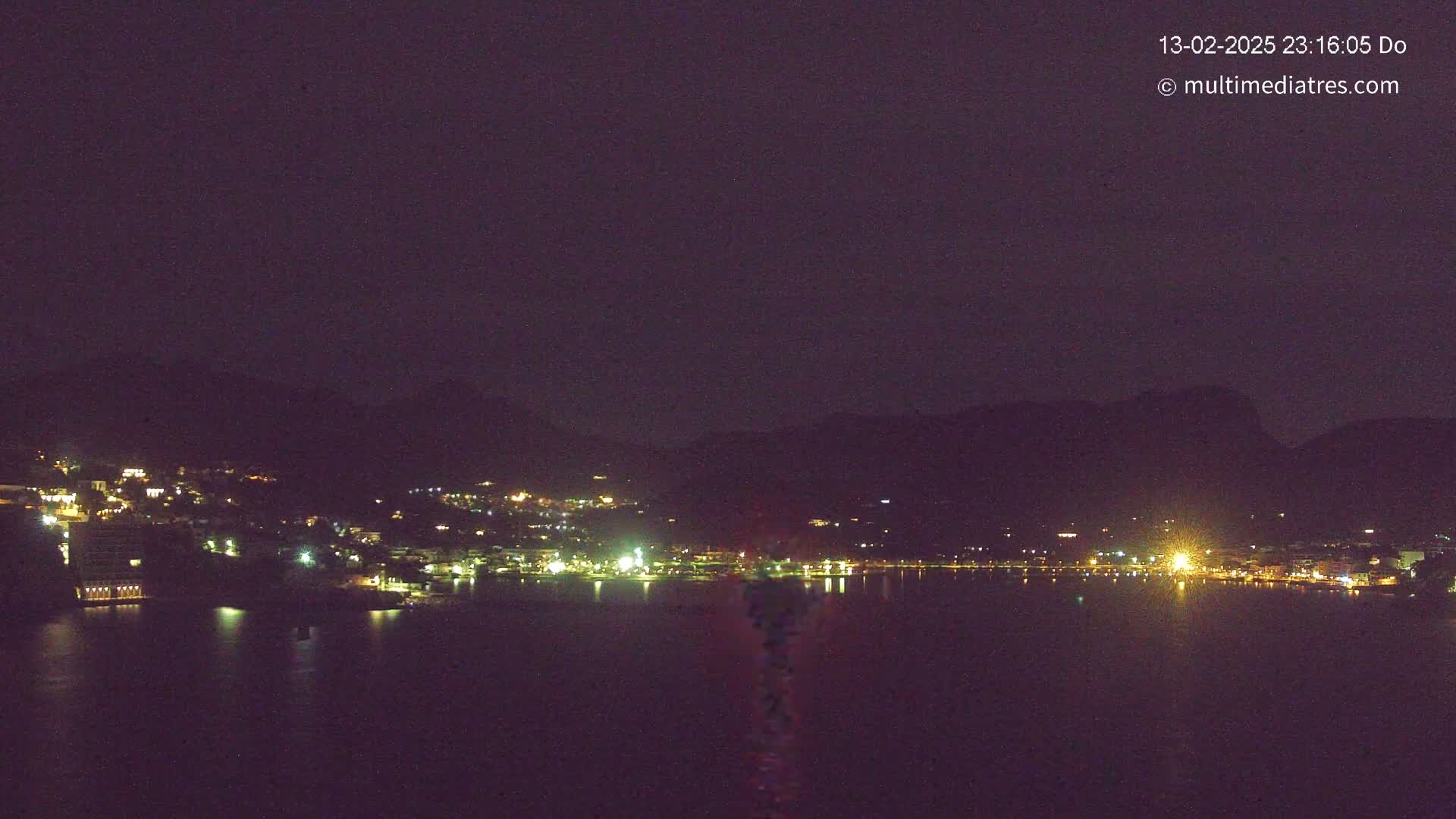 A nighttime view of a town's lights along a dark body of water, nestled against a mountain range under a clear, dark sky.