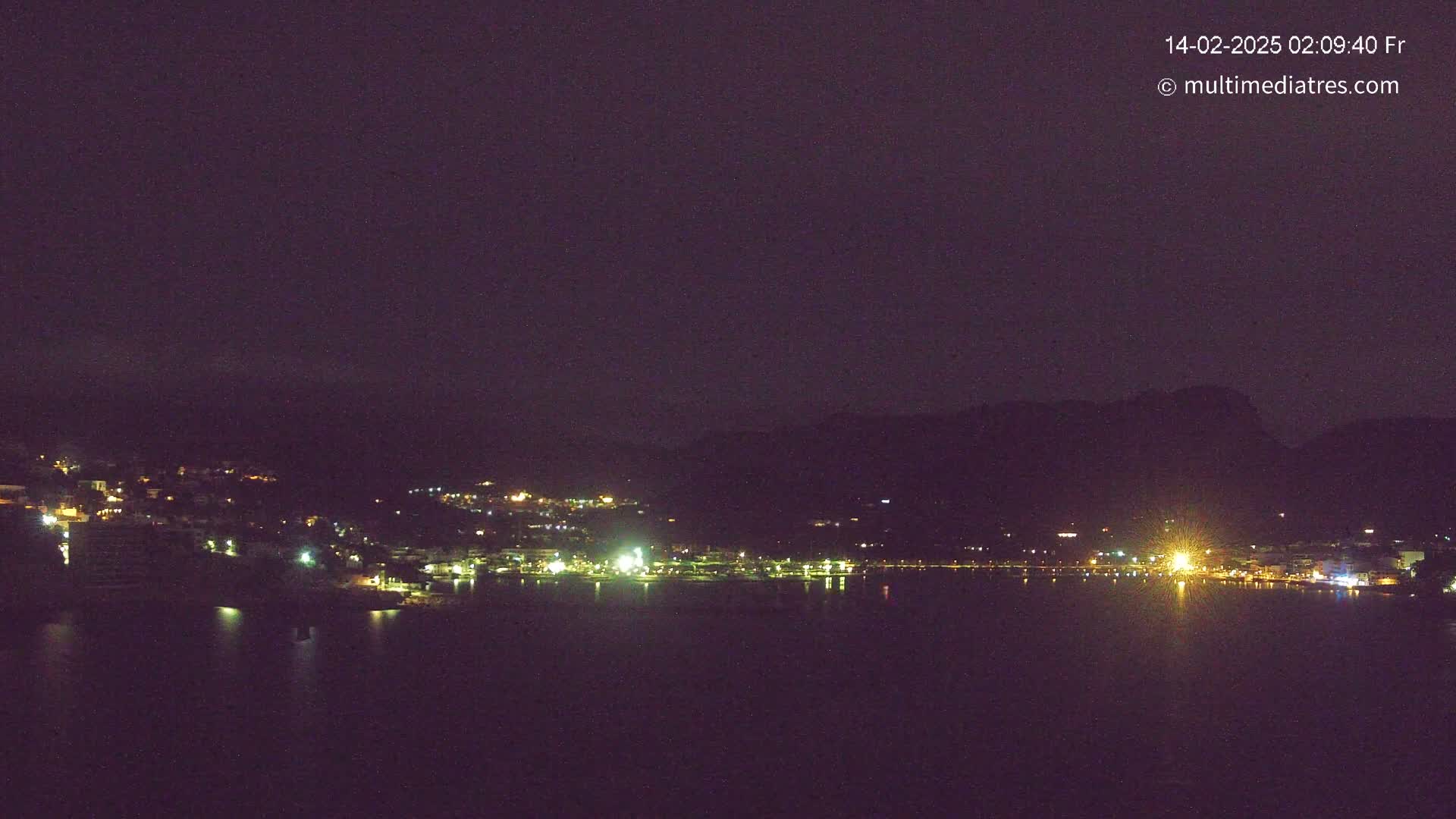 A nighttime view shows a dark body of water reflecting the lights of a town nestled against dark hills under a dark, clear sky.