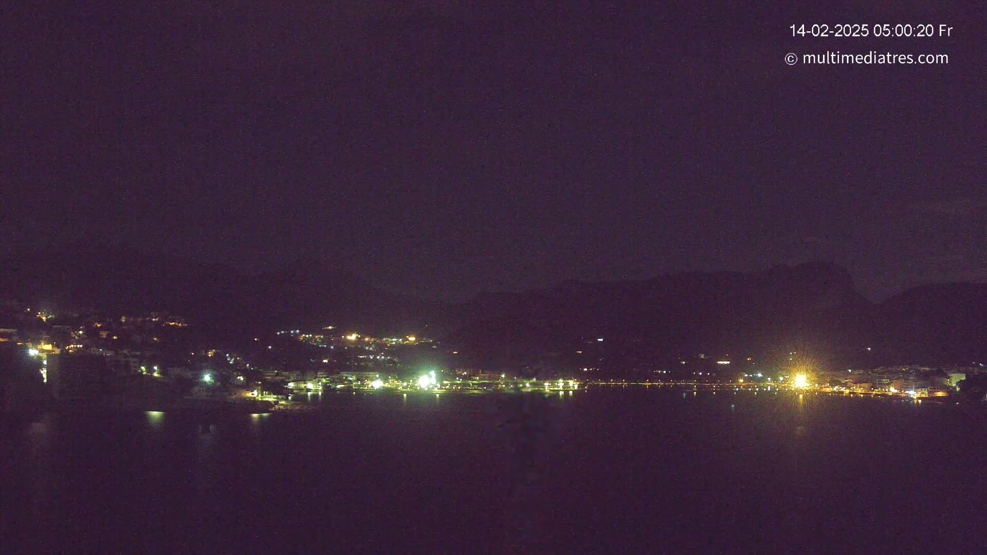 A nighttime long shot shows a dark, calm body of water reflecting the scattered lights of a town nestled against dark hills under a clear, dark sky.
