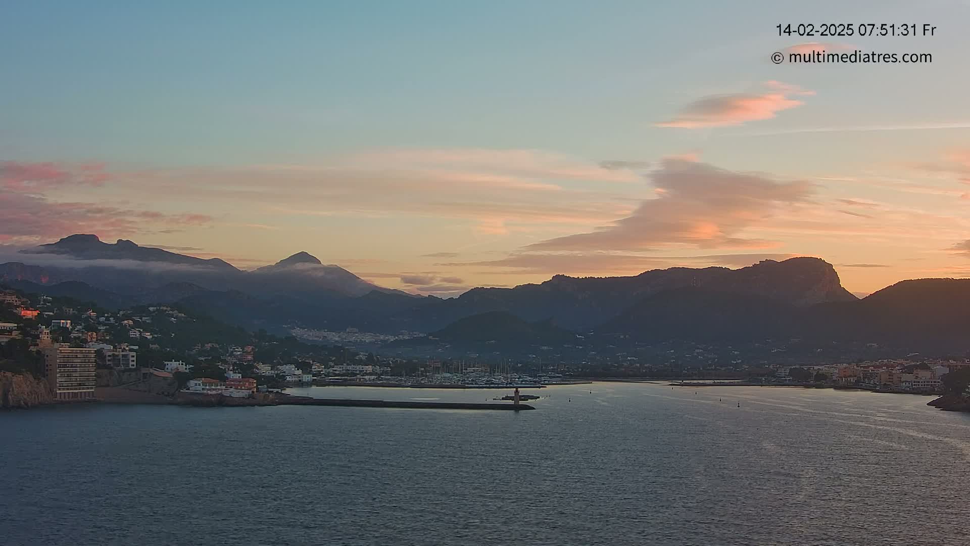 A coastal town nestled at the base of mountains is bathed in the soft light of a sunrise with a mostly clear sky showing streaks of pink and orange.