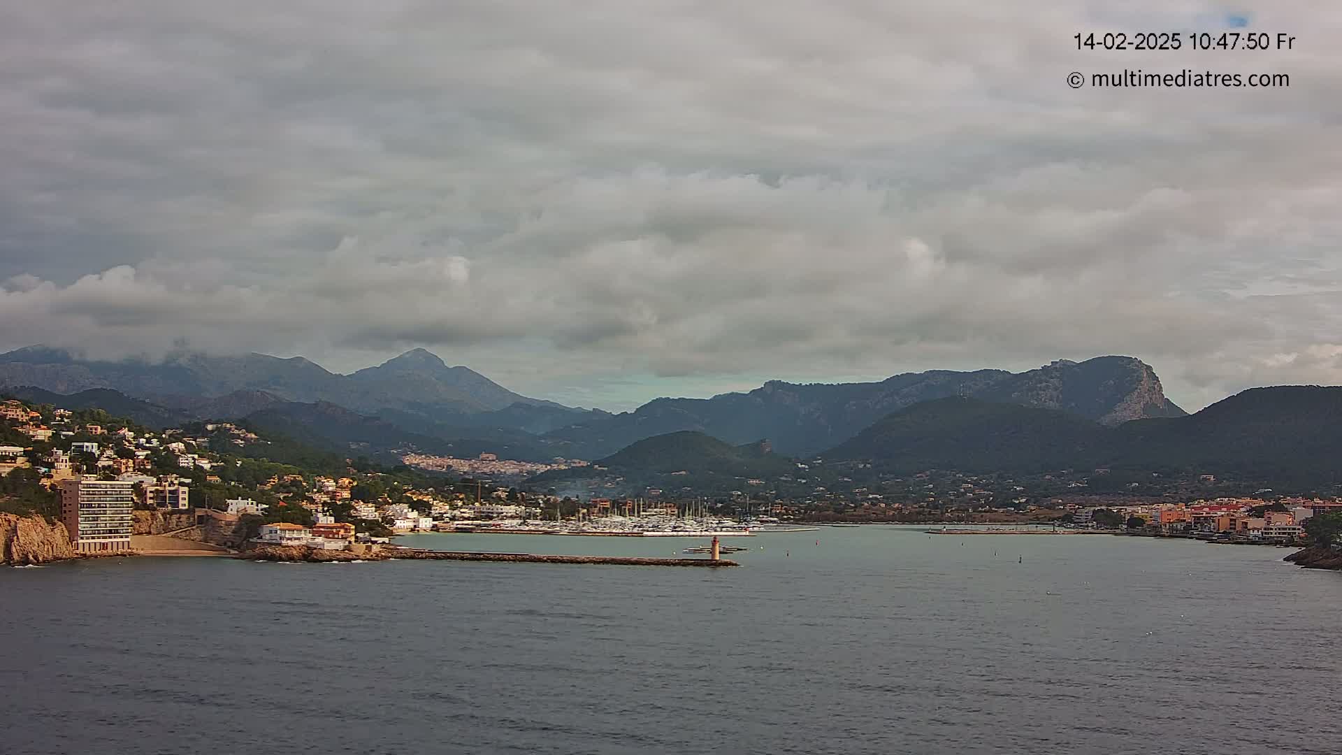 A coastal town nestled between dark mountains under a mostly cloudy sky sits on a calm body of water with a marina filled with boats.