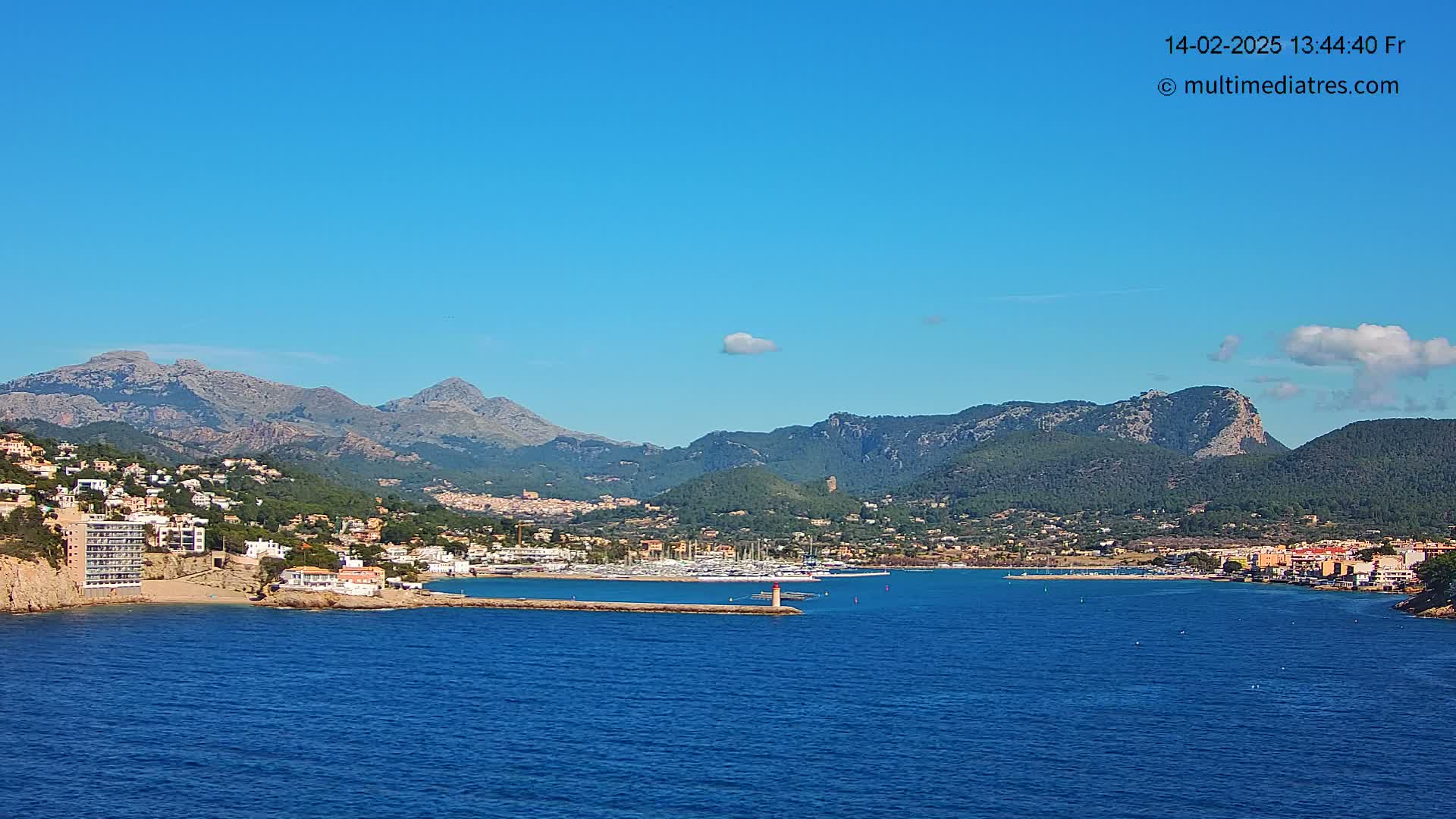 A coastal town nestled against a backdrop of mountains under a clear blue sky with a few scattered clouds.
