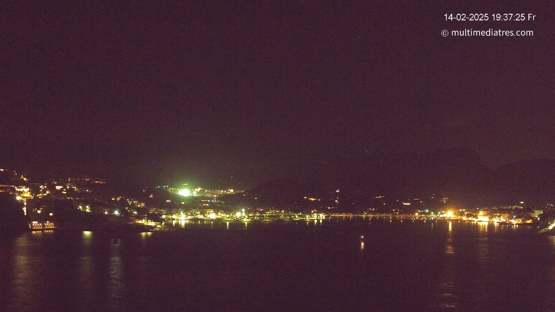 A nighttime view of a town's lights scattered along a dark shoreline and hills, reflected in a calm body of water under a clear, dark sky.