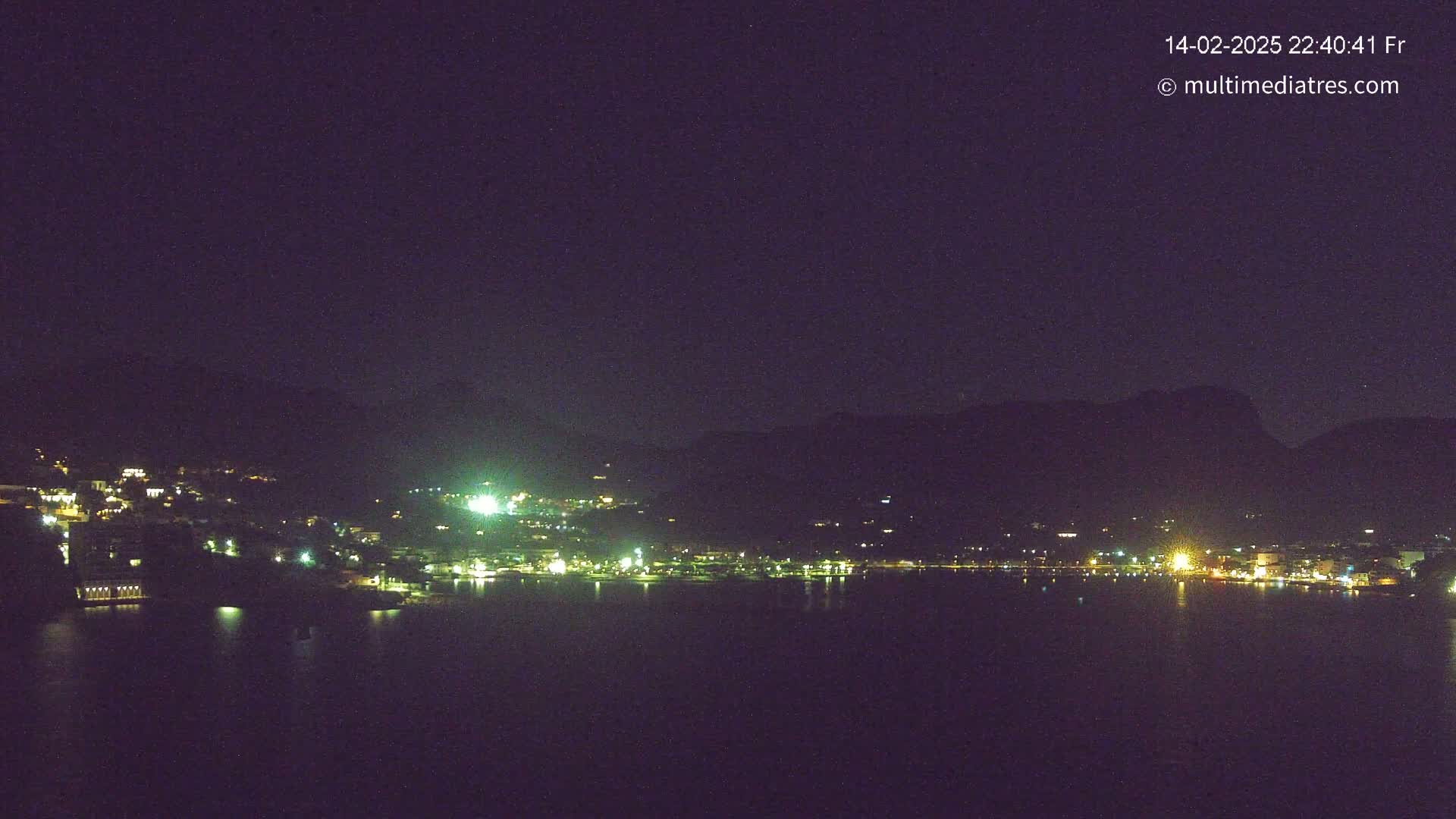 A nighttime view of a town nestled beside a body of water, illuminated by scattered lights against a dark, clear sky and silhouetted mountains.