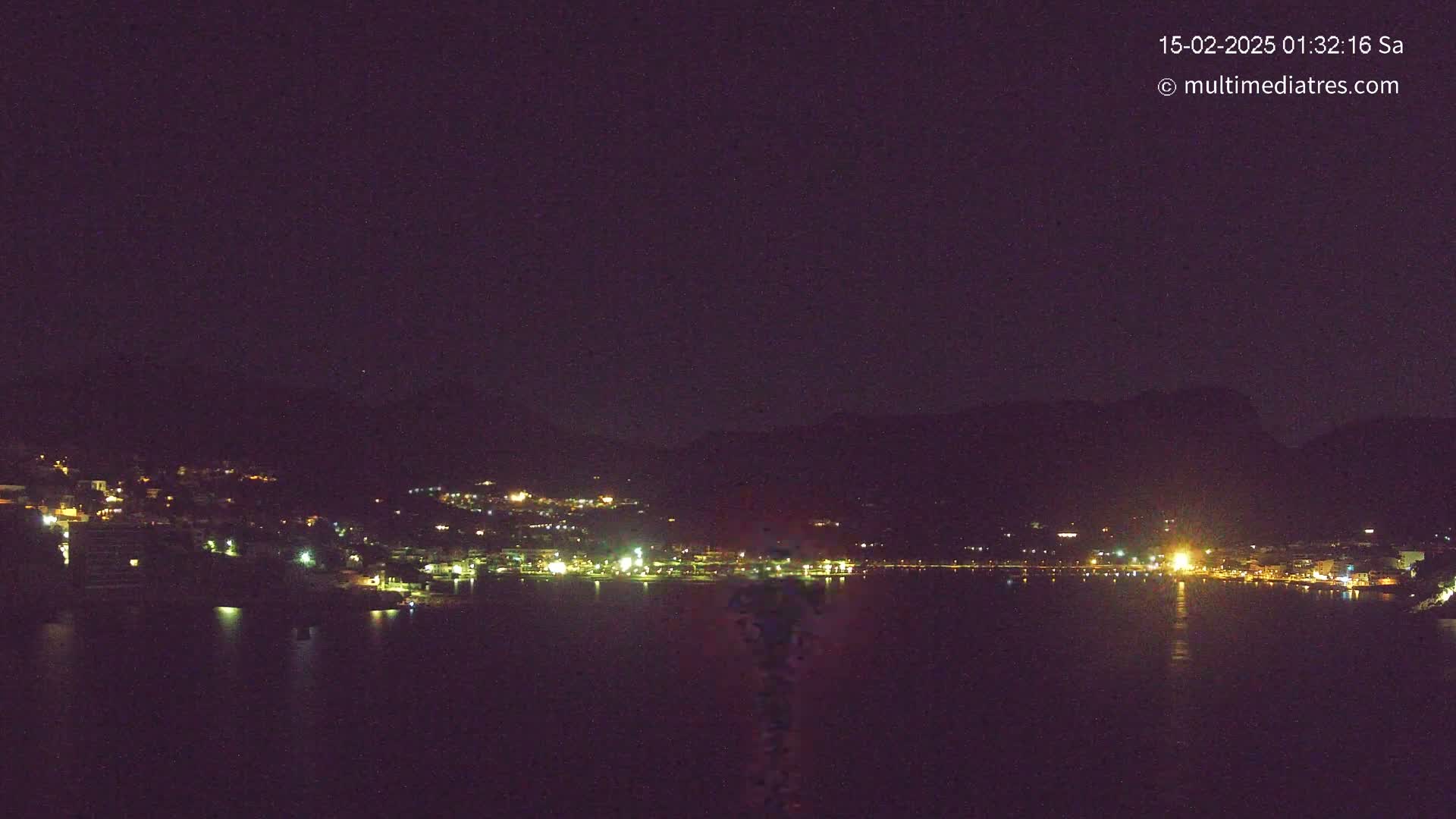 A nighttime view of a town's lights across a dark body of water, nestled against a mountainous backdrop under a clear, dark sky.
