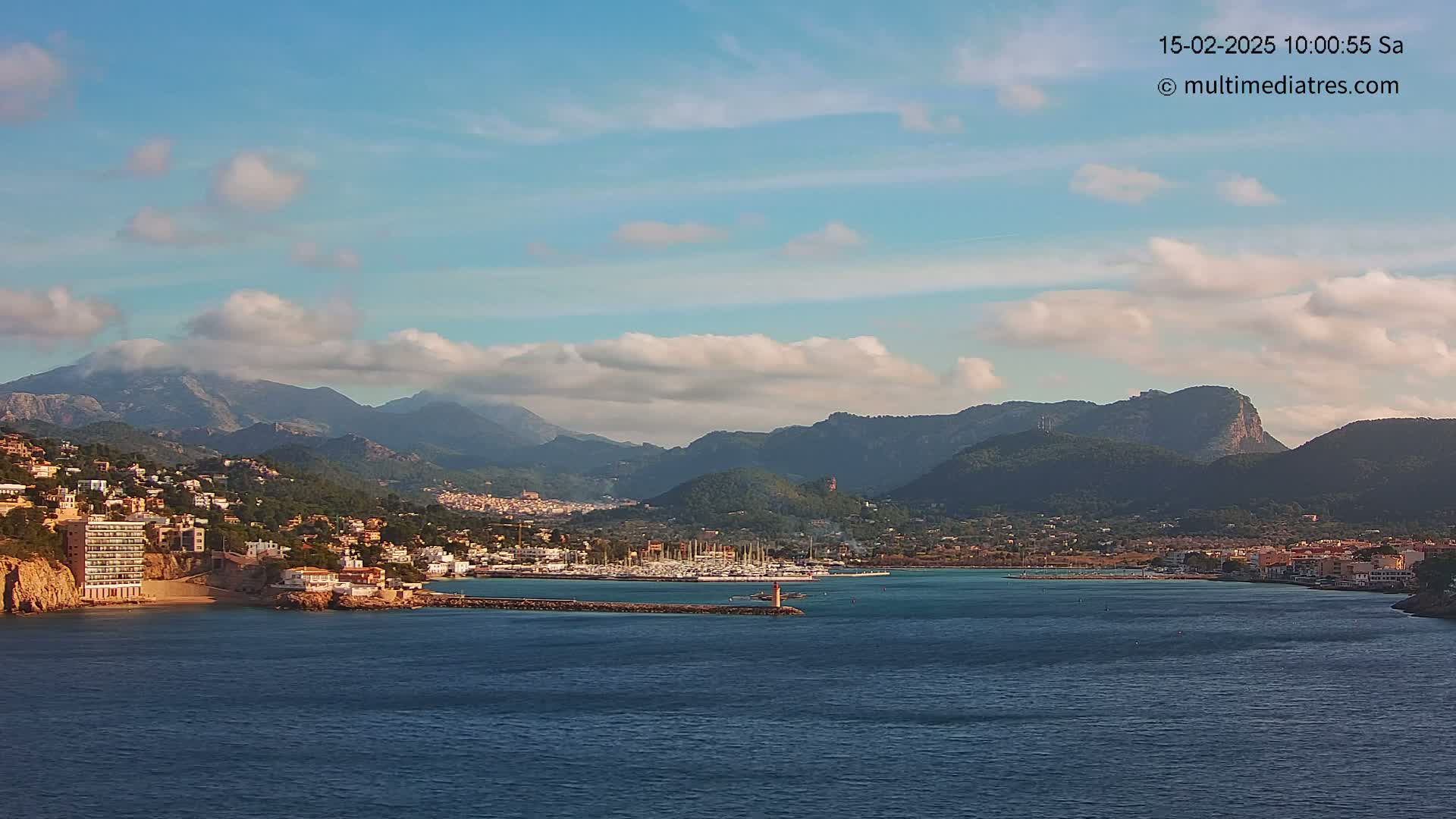 A coastal town nestled against a mountain range under a partly cloudy sky, with a marina filled with boats in the foreground.