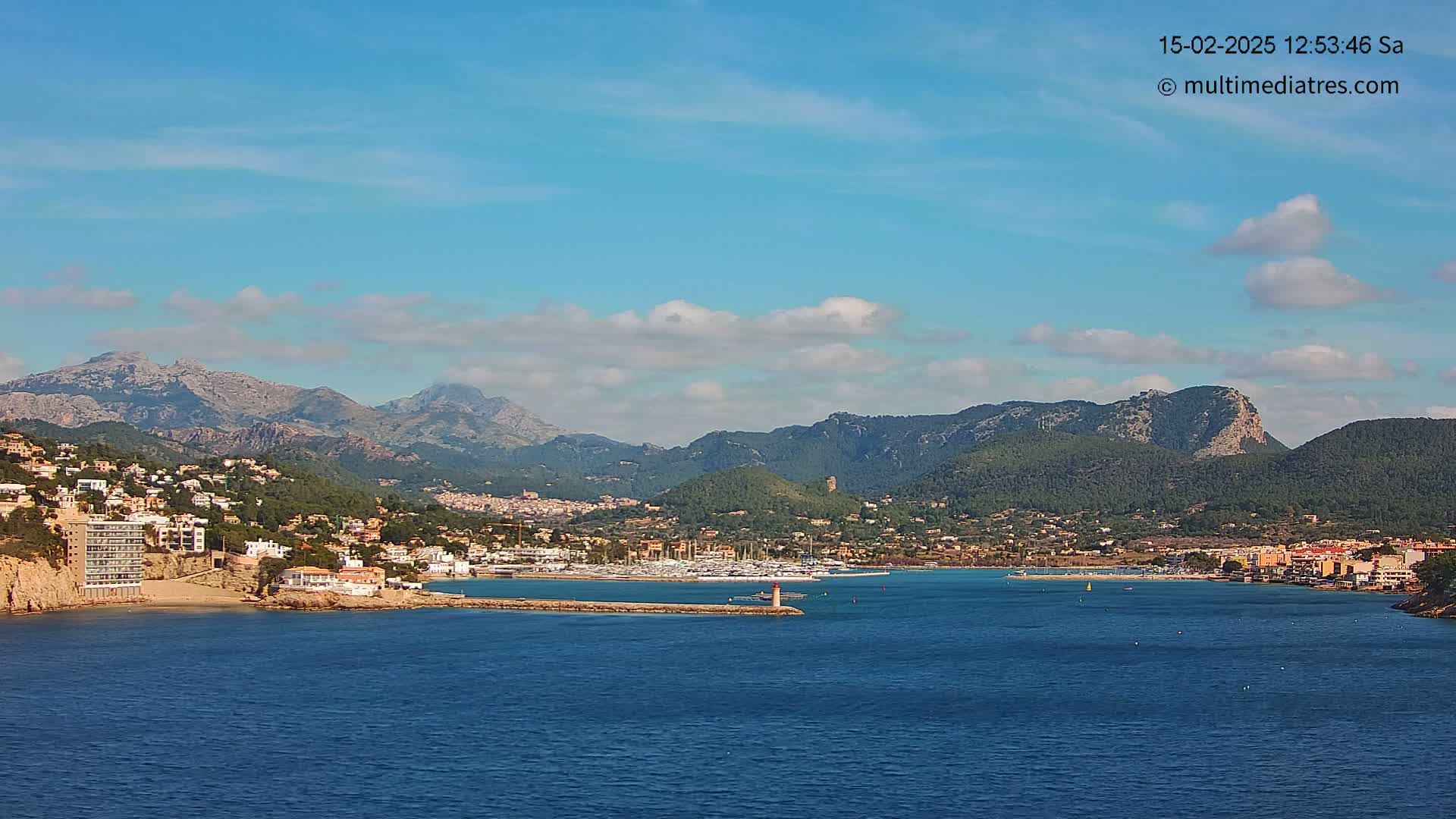 A coastal town nestled between mountains and a calm blue sea under a mostly sunny sky with a few scattered clouds.