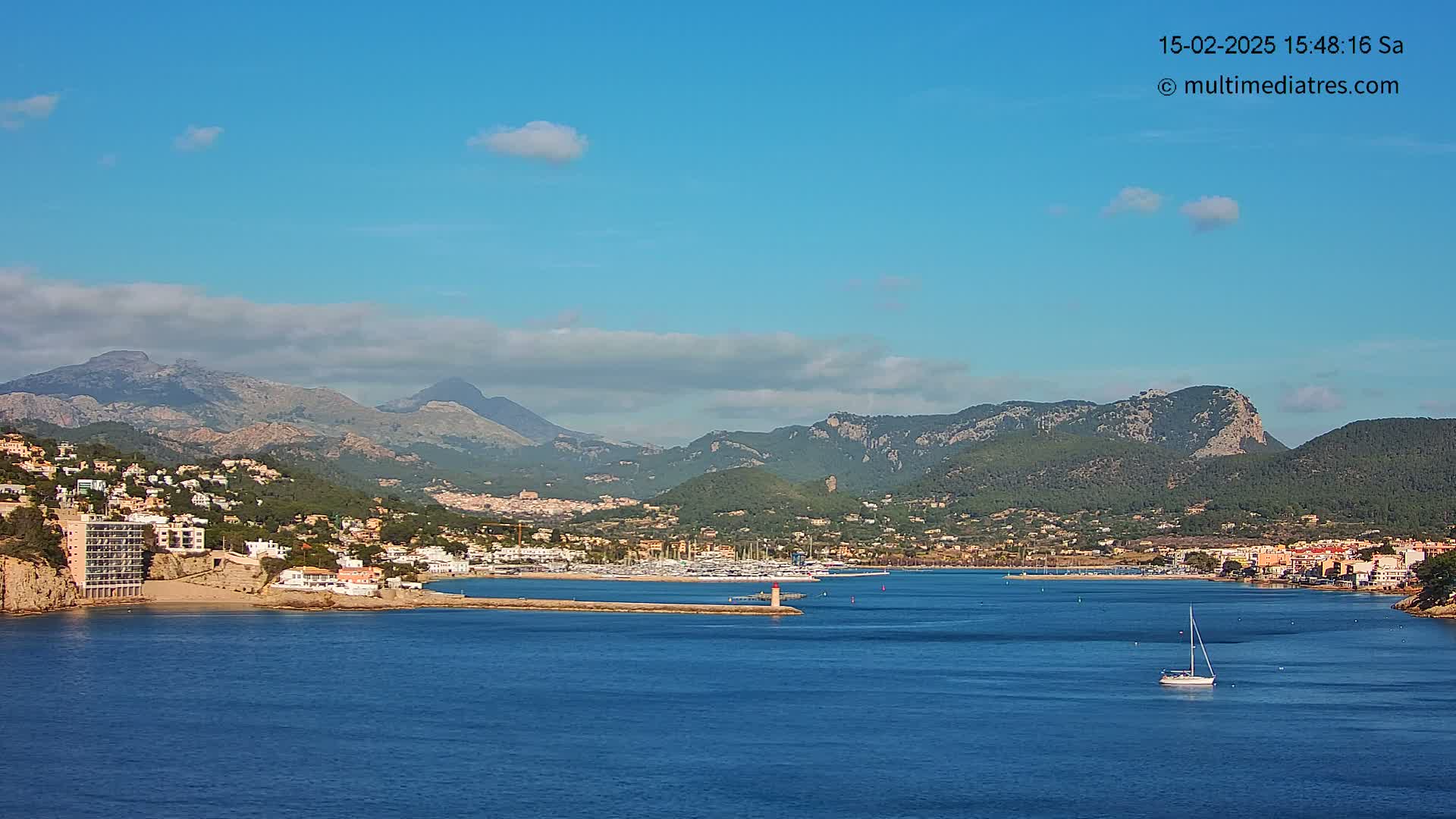 A sailboat is visible in a calm bay, nestled between a coastal town and a mountain range under a mostly clear blue sky with a few scattered clouds.