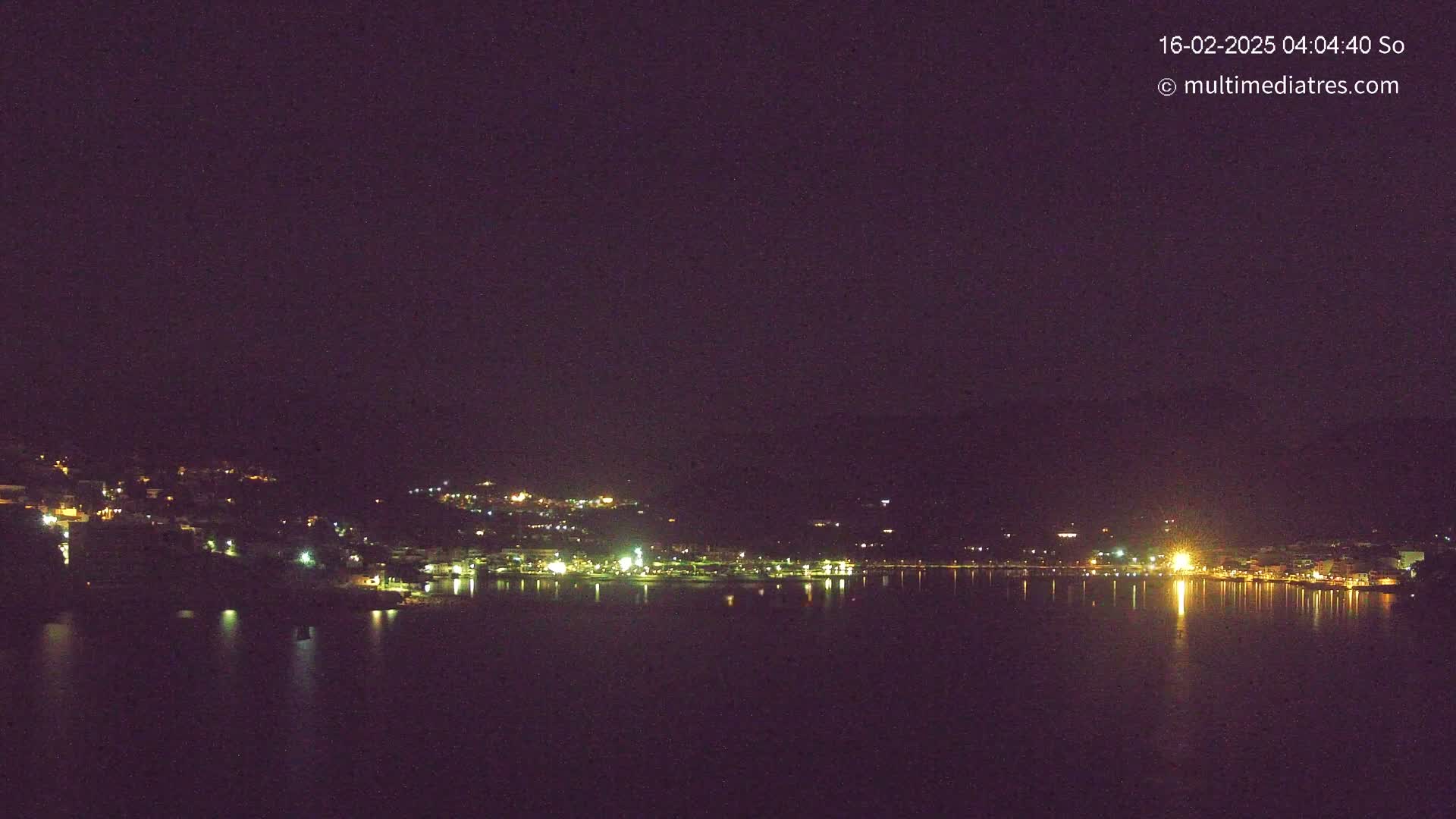 A nighttime view of a town situated on a body of water, with its lights reflecting on the calm water under a dark sky.